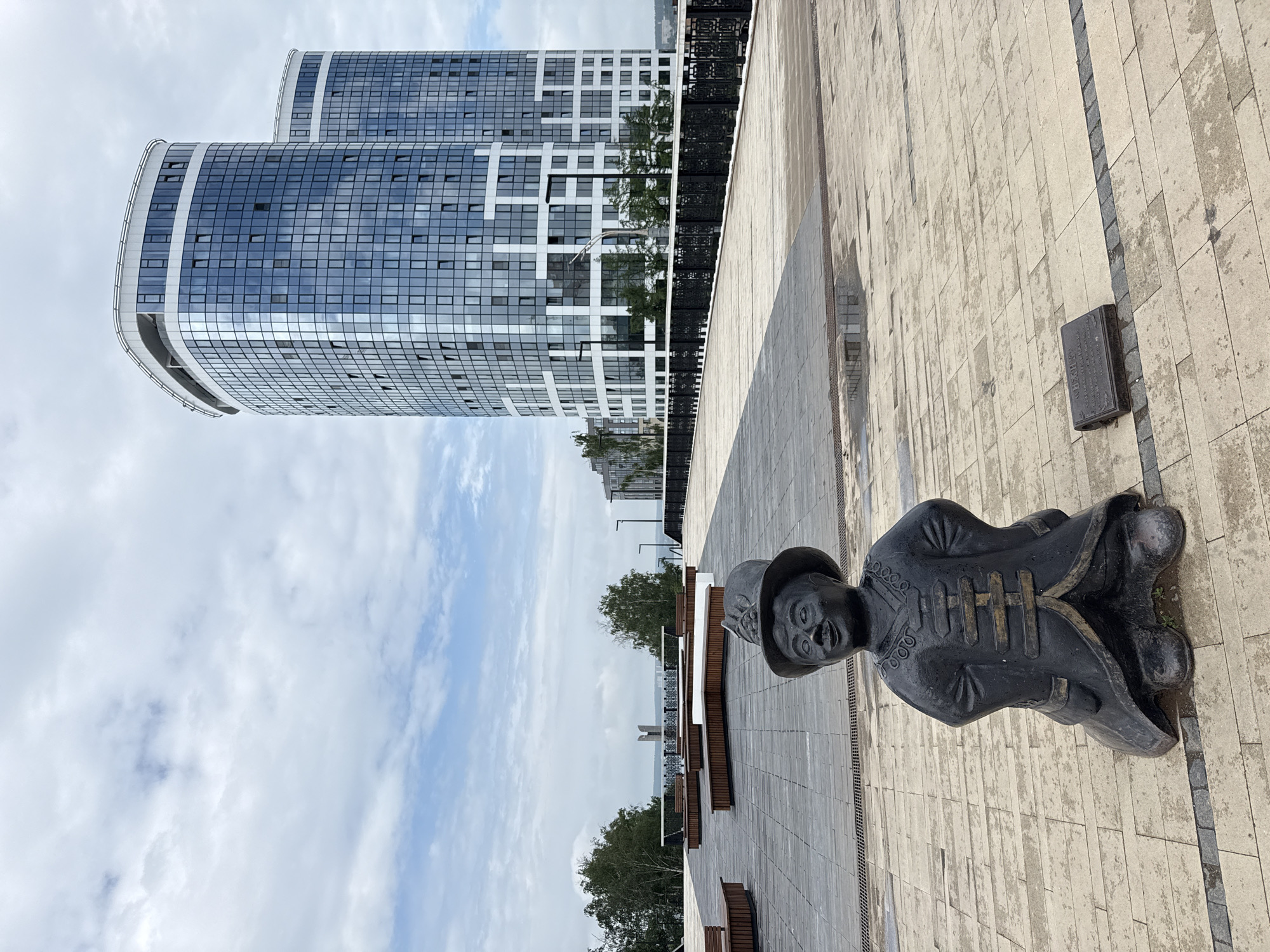 This is a wide shot of a modern waterfront promenade in Izhevsk, Russia. In the foreground, a small, dark bronze statue of a man wearing a traditional hat and ornate coat stands on light-colored paving stones. A small bronze plaque is visible on the ground to the right of the statue. Beyond the statue, the promenade features sleek, modern benches and a black metal railing. In the background, two towering, curved glass-fronted buildings rise against a cloudy sky. Trees and a distant bridge are visible on the horizon. The ground appears wet in places, suggesting recent rain.