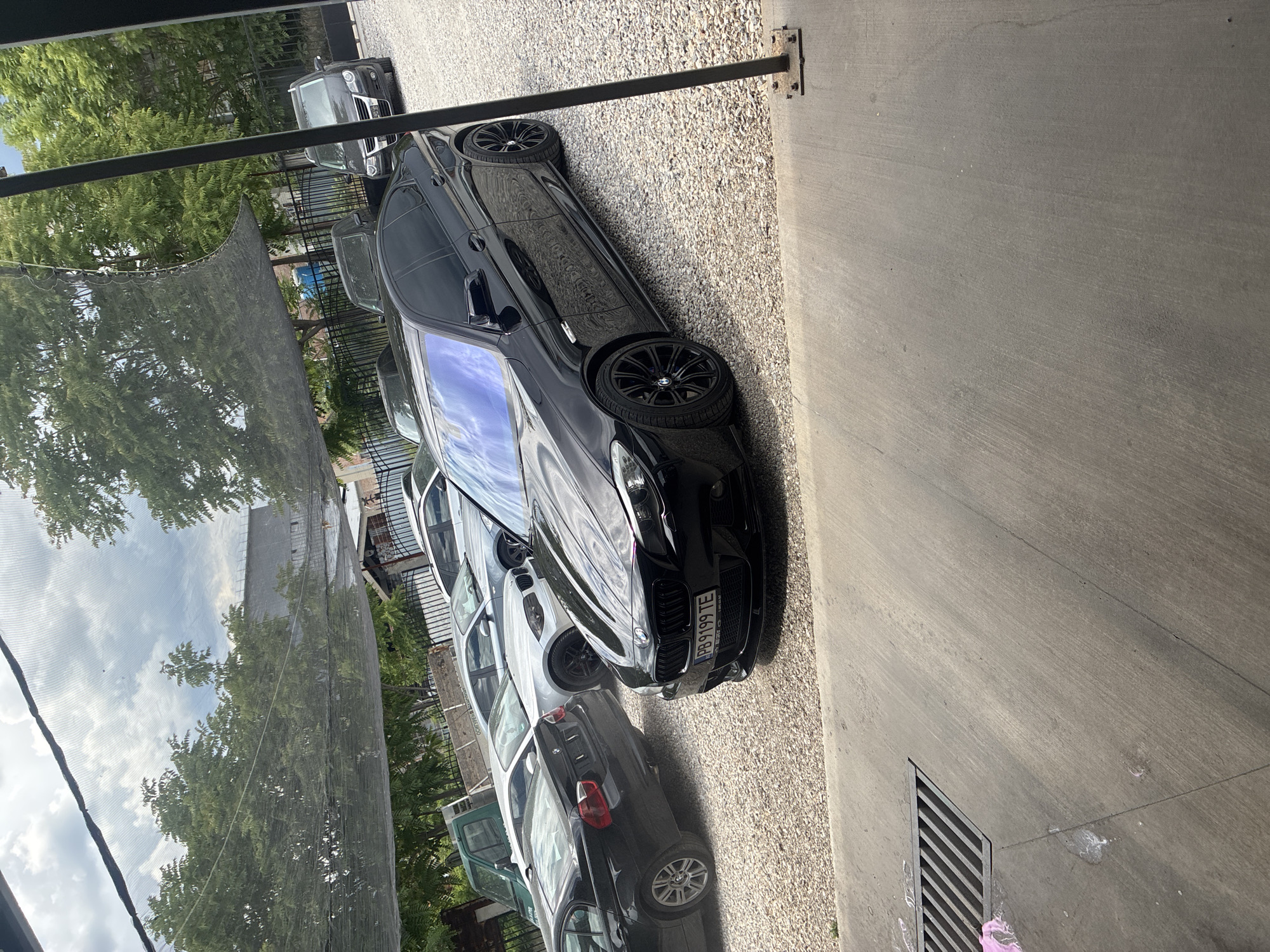 The image shows a row of parked cars along a gravel driveway next to a concrete surface in an urban or suburban area. The cars vary in model and color, including black, white, and green. Trees and a netting structure are visible in the background. A drain is present in the concrete surface. The photo seems to be taken from a slightly elevated position. The license plate of the closest black car reads "PB 9199 TE." The city and specific location are unknown.