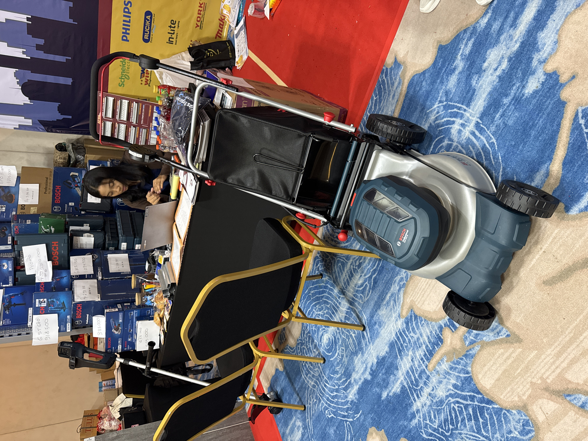 Here is a concise description of the image:
The image shows an indoor sales event, possibly a trade show or promotional event in Denpasar, Indonesia, indicated by the text on the product boxes (including price tags with Indonesian Rupiah values). A woman is sitting at a table, possibly working on a laptop, surrounded by boxes of Bosch power tools and other products from brands like Philips and Schneider Electric. A lawnmower sits to the right of the table on a blue patterned rug. There are chairs stacked to the left of the table.