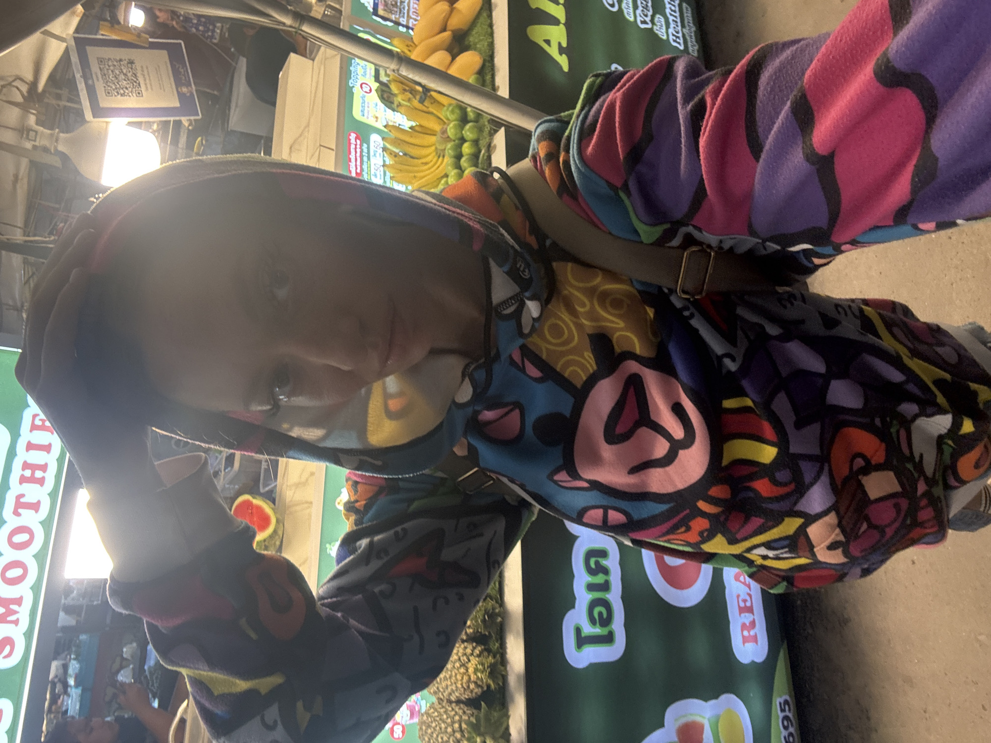 The image shows a person taking a selfie or photo at a brightly lit fruit or smoothie stall, likely in an evening market setting in บ้านบางรักษ์, Thailand. The subject wears a distinctive, colorful hoodie featuring a vibrant, abstract pattern with cartoon-like characters in shades of purple, pink, blue, yellow, orange, red, and black. A khaki shoulder bag strap is visible across their chest. Bright artificial lights illuminate the scene, causing glare on the person's face, which is partly obscured, showing a neutral expression.
In the background, various fresh fruits are displayed on the stall, including bunches of yellow bananas, green limes or small citrus fruits, and yellow mangoes. Pineapples and watermelon slices can also be seen. Multiple signs are present. A prominent green sign on the left displays "SMOOTHIE" in white letters. Another green sign features Thai script and the English word "REAL," along with numbers like "60" and "595," likely indicating prices. Additional small signs show "Topping" and "50," and faint text suggesting "Vegan" and "Health." A QR code is visible on a white sign with accompanying Thai text. The scene depicts a casual interaction at a market stall, characterized by vivid colors and artificial lighting.