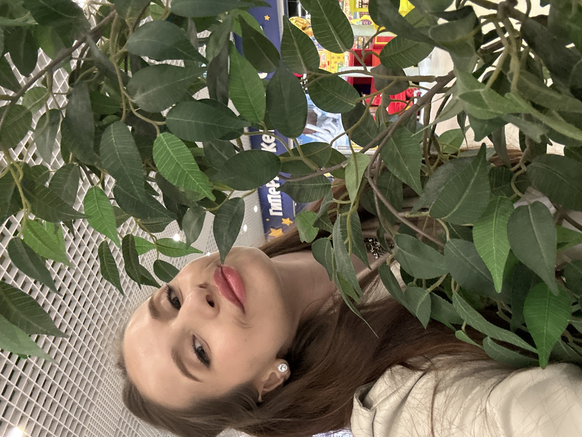 The image shows a woman posing behind artificial foliage, with a retail setting visible in the background. The woman has light skin, brown hair, and is wearing a light-colored jacket. She is wearing earrings. Part of her face is visible through the artificial leaves. The background reveals store shelving with various products. Some text in Cyrillic script is present on store signage. A white grid ceiling is visible on the left of the image. It appears to be an indoor setting. The exact location in Yekaterinburg, Russia, cannot be determined without further information.