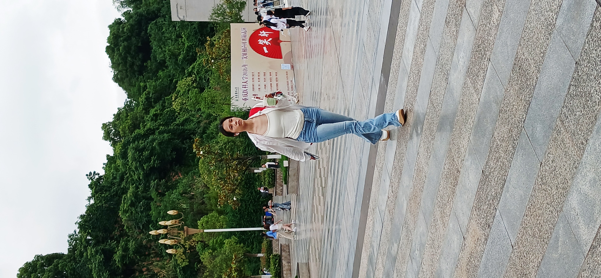 A woman is walking on a paved walkway in Chongqing High-tech Zone, China. She has dark hair pulled back, and wears a white tank top, light blue jeans, and a white shirt. She is holding a mobile phone in one hand, and a cup in the other. Behind her is a sign with Chinese characters and a red fan design, as well as green trees and foliage on a hill. Other people are visible in the background. The walkway is made of long, rectangular paving stones in light gray and tan colors.