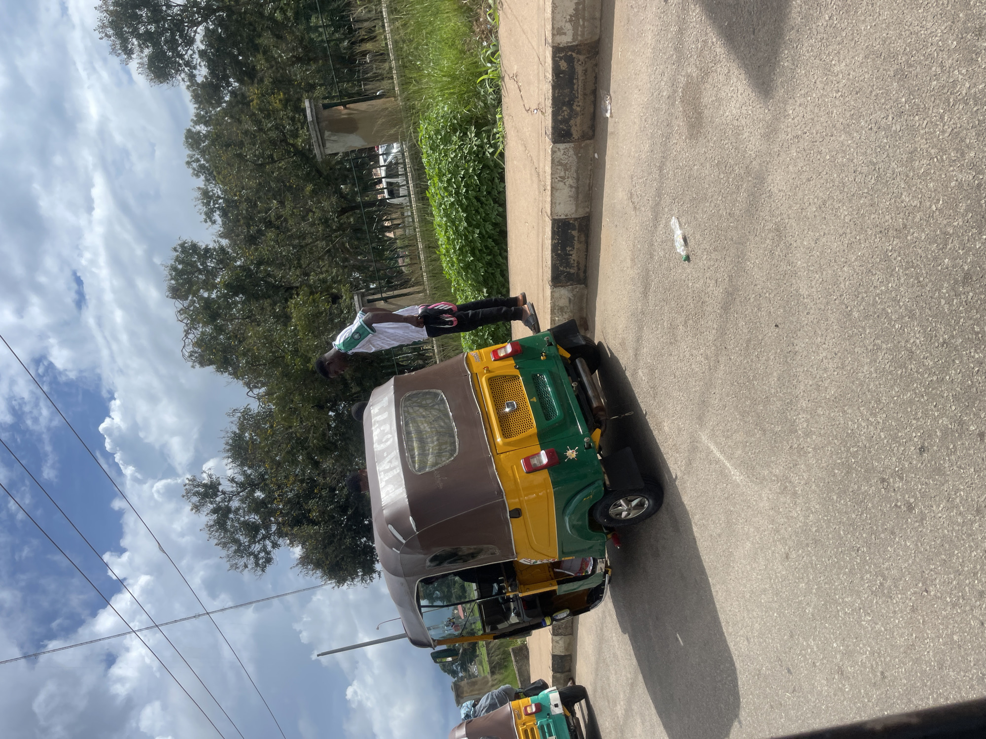 A yellow and green auto-rickshaw is parked on the right side of a paved road. A man wearing a white and green shirt and black pants stands on the sidewalk to the right of the auto-rickshaw, facing it. Another auto-rickshaw is partially visible on the left side of the frame. Trees line the background behind a black metal fence. The sky is partly cloudy. There is a plastic bottle on the road. The scene is in Bar Jirgi Summa, Nigeria.