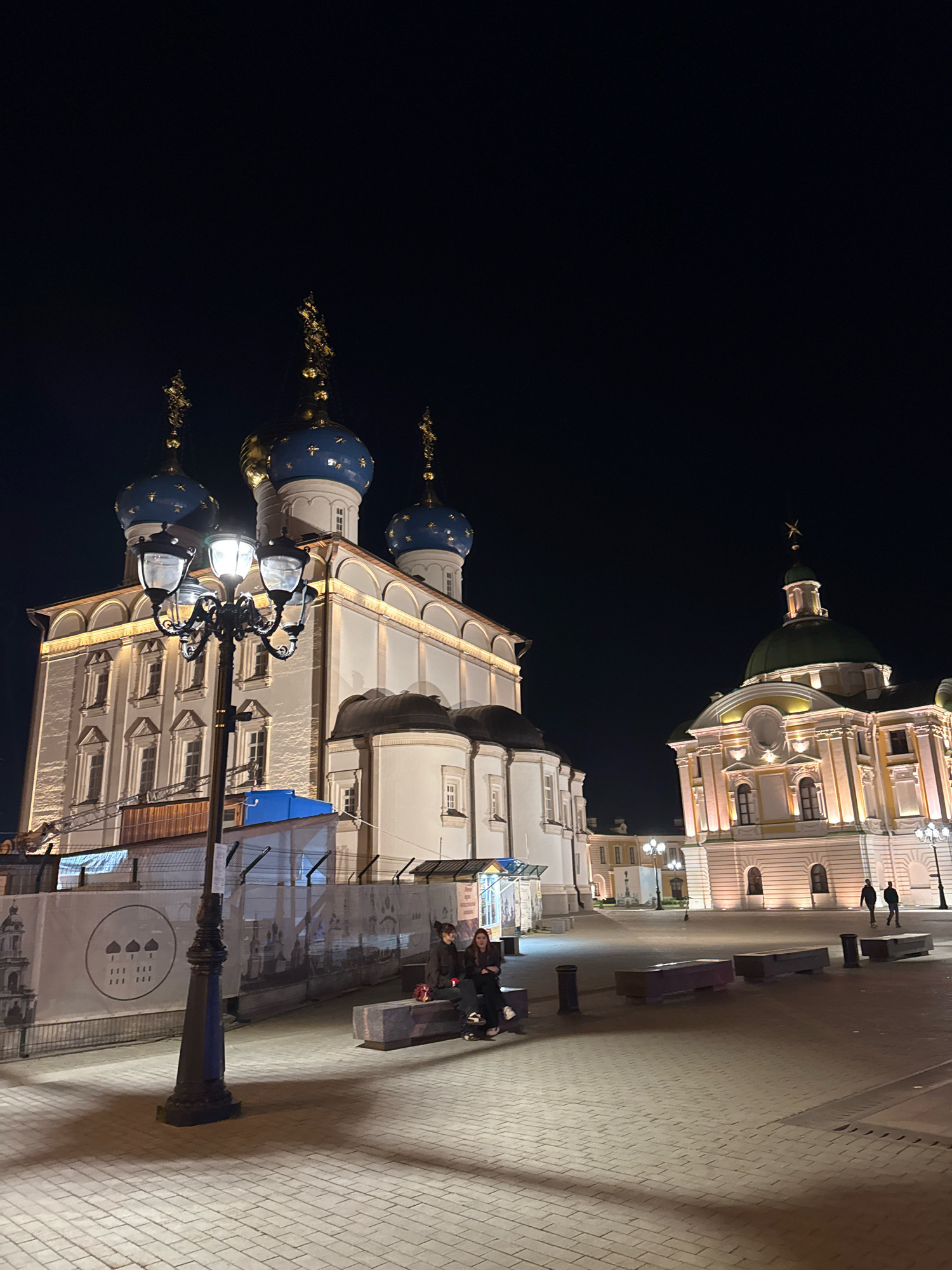 The image depicts a night scene in Tver, Russia, likely on the territory of the Ascension Cathedral. In the foreground, a black street lamp with four lights stands prominently. To the left, a large white building with distinctive blue domes topped with golden spires dominates the frame. This building is illuminated from below, highlighting its architectural details and numerous windows. To the right, a smaller, ornate building with a green dome and golden star is visible, also brightly lit. The ground is paved with light-colored bricks. Two women are seated on a long, dark bench in the middle ground, facing away from the camera. In the background, a large banner with architectural illustrations and text is displayed, and a few other figures are walking in the distance. The sky is dark, indicating it is nighttime.