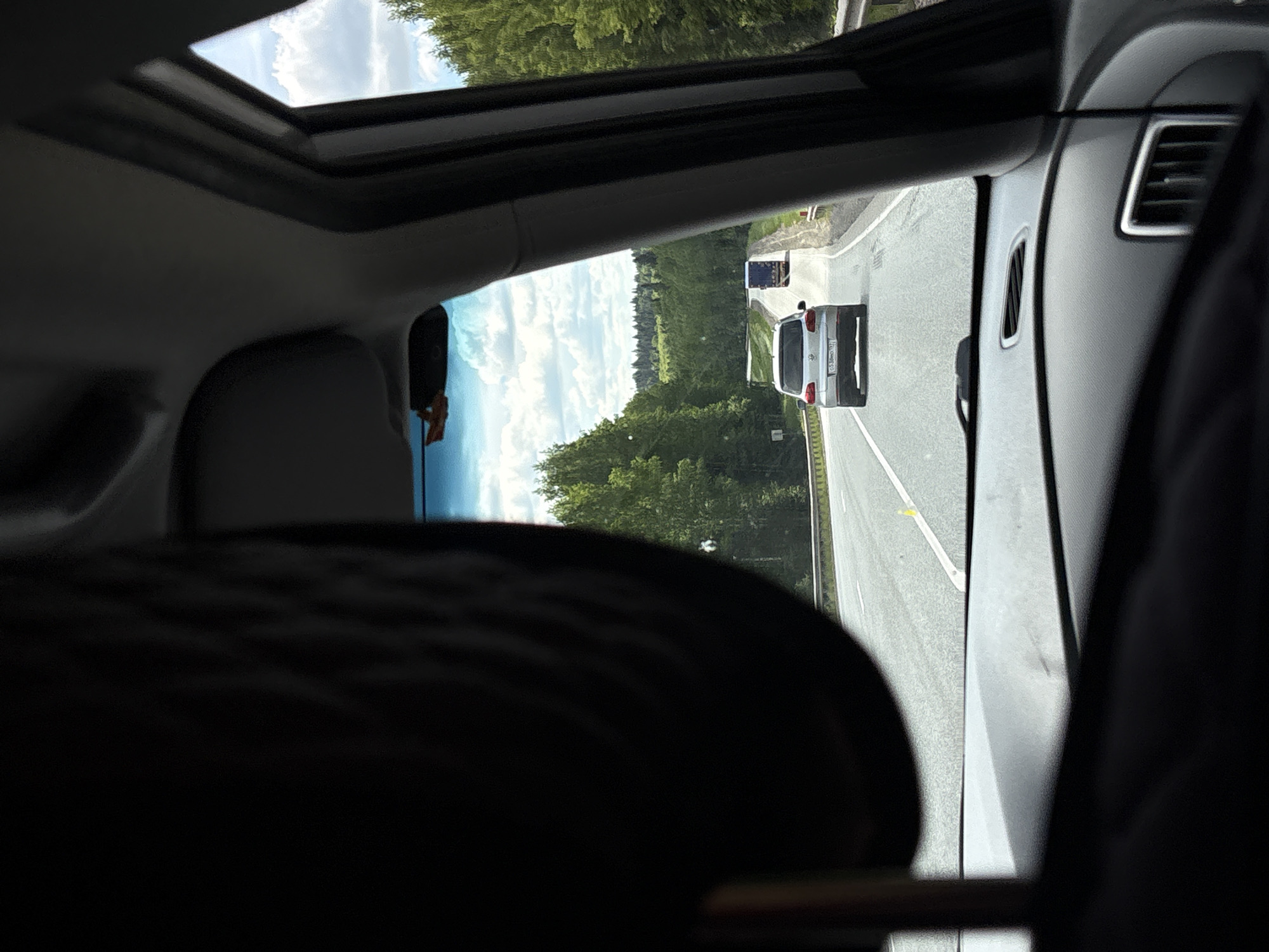 The image presents a forward-facing view from inside a vehicle on a multi-lane asphalt highway, located in the vicinity of the city of Verkhnyaya Talitsa, Russia. A dark headrest occupies the left foreground, while the vehicle's dashboard, right A-pillar, and rearview mirror (from which a small, possibly orange-colored object hangs) partially frame the scene. The highway features visible lane markings and is flanked on both sides by dense green deciduous trees, suggesting a rural or forested environment. Guardrails line the road's edges. The sky above is partly cloudy with scattered white cumulus clouds. Ahead, a silver Volkswagen Tiguan SUV, bearing the Russian license plate "O 530MH 197" (Moscow region), is visible traveling in the same direction. Further down the road and appearing to ascend a gentle incline, a blue semi-truck is observed, with another lighter-colored vehicle faintly visible beyond it. No individuals are discernible within the frame.