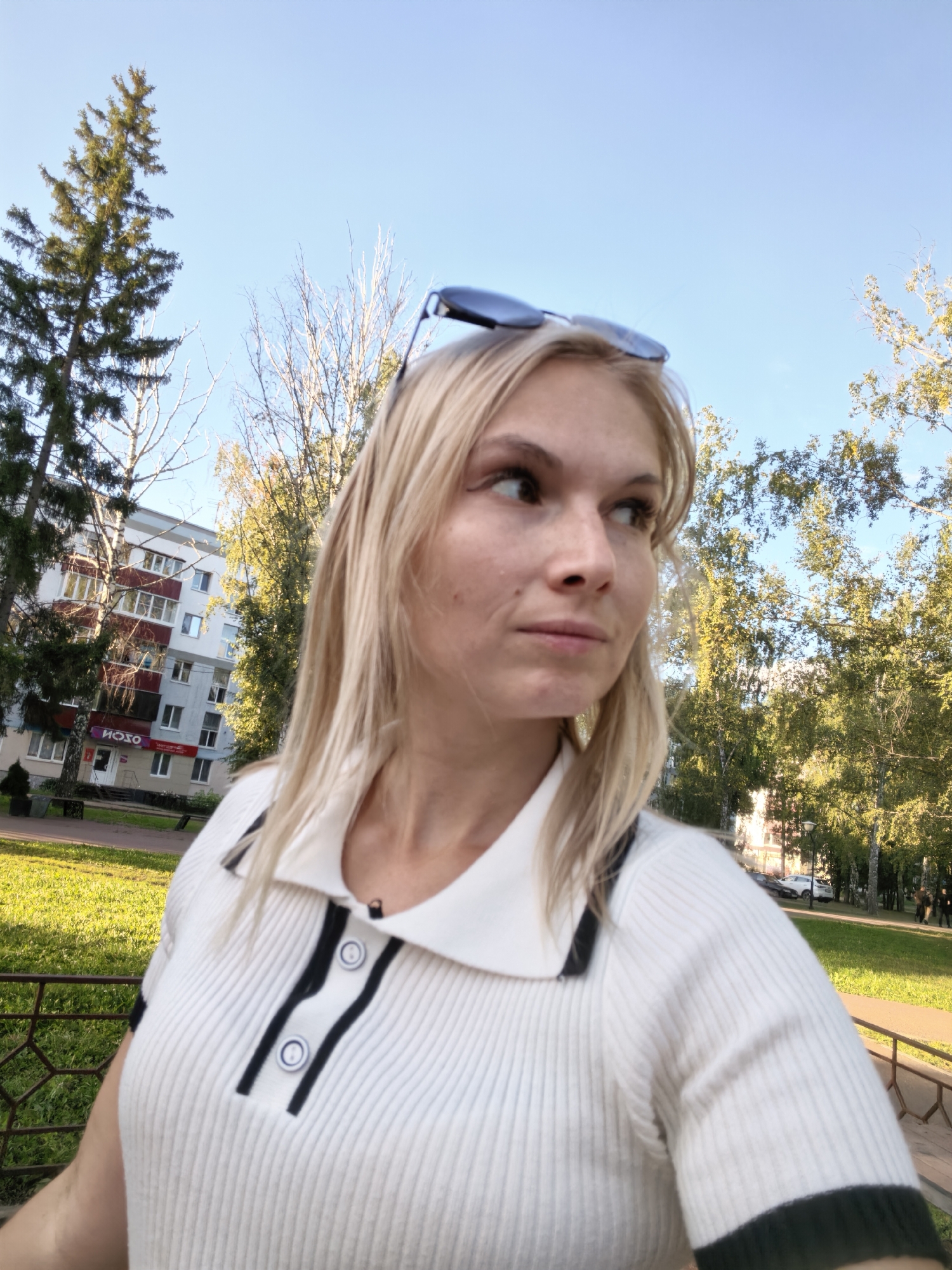 A blonde woman with sunglasses perched on her head is captured in a close-up, selfie-style shot outdoors. She is wearing a white ribbed polo shirt with black trim and buttons. The background reveals a residential building with balconies, trees with green and yellow foliage, and a clear blue sky. The environment suggests a park or residential area in Sterlitamak, Russia, with sunlight illuminating the scene. There are no other visible people or discernible actions.