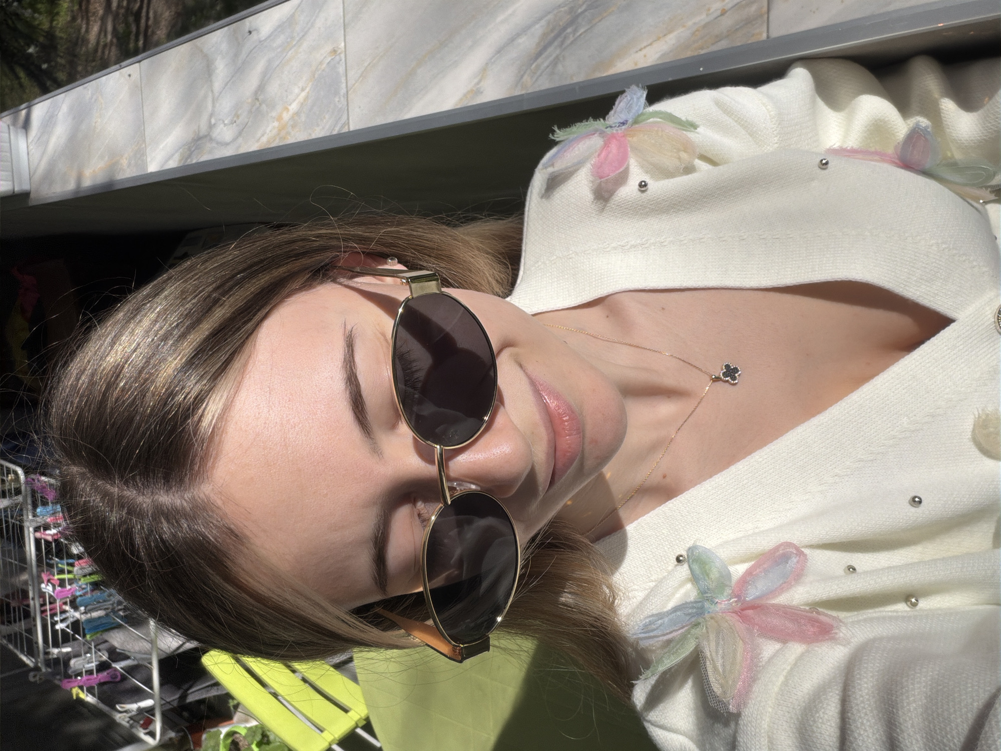 A woman is posing outdoors in sunlight. She is wearing sunglasses and a white, flower-decorated cardigan. A delicate gold necklace with a four-leaf clover pendant is visible on her neck. The background includes a stone wall and a green chair. Part of a metal clothes drying rack is also visible in the background. There's insufficient data to determine the exact location in the city of Plovdiv, Bulgaria.