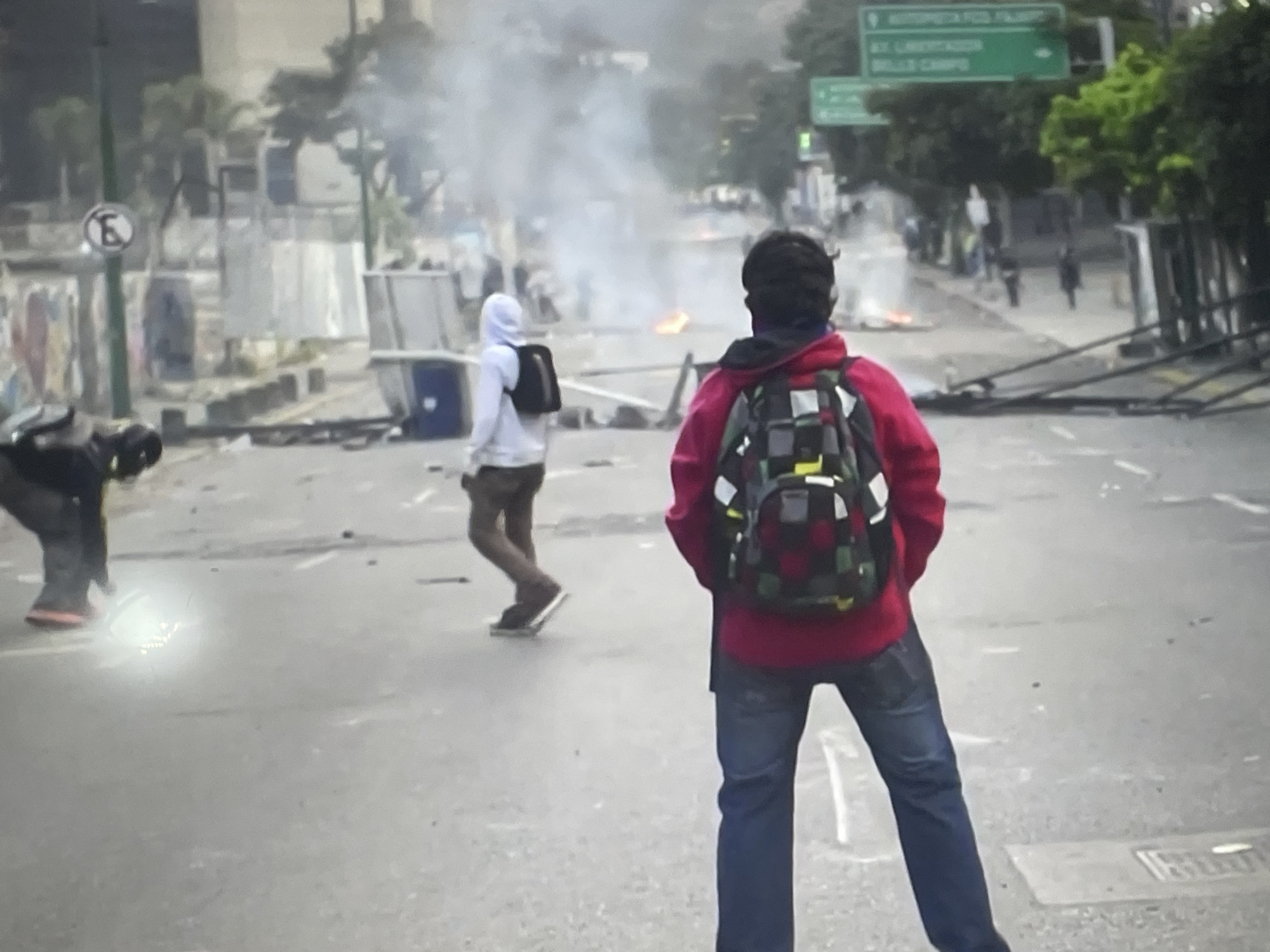 The image depicts a street scene, potentially a protest or riot, in Unknown, Unknown. Three individuals are visible: one on the left, obscured and crouching; another in the center, walking with a backpack and wearing a white hooded sweatshirt; and a third, closer to the foreground, facing away from the viewer, wearing a red jacket, jeans, and a backpack. Smoke rises in the background, suggesting burning debris. A sign on the right indicates "Alt. Libertador FCO. Fajardo Bello Campo". A "no parking" sign is visible on the left side of the road. Barricades and scattered debris litter the street.