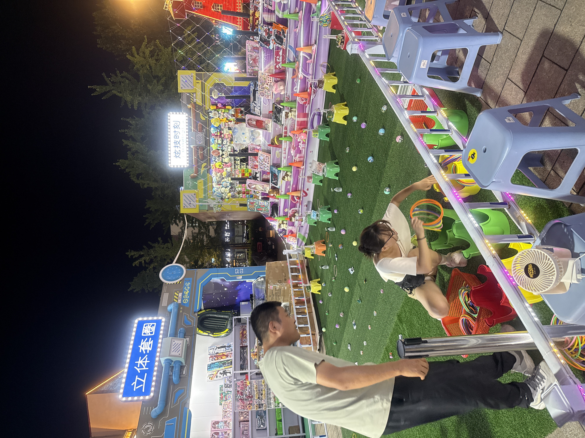 This image captures a nighttime scene at an outdoor fair or carnival, possibly in Qinhuangdao, China. In the foreground, a man in a light-colored t-shirt and dark pants stands to the left, observing the activity. To his right, a woman in a white t-shirt and dark shorts is crouched down, reaching into a red basket filled with colorful hoops. She appears to be participating in a ring toss game.
The game area is covered in artificial green turf, scattered with small, colorful balls and various game prizes. In the background, a brightly lit stall displays an assortment of stuffed animals and other carnival prizes. Above the stall, two illuminated signs are visible: one in blue reads "立体套圈" (lìtǐ tào quān), which translates to "3D Ring Toss," and another smaller sign with Chinese characters, likely indicating the name of the game or event. Several green and yellow plastic stands with buckets are set up on the turf, serving as targets for the ring toss game. Blue plastic stools are arranged for spectators. A small, beige fan is placed on a railing in the lower right corner. The overall atmosphere is festive, illuminated by the bright lights of the stalls and decorative string lights.