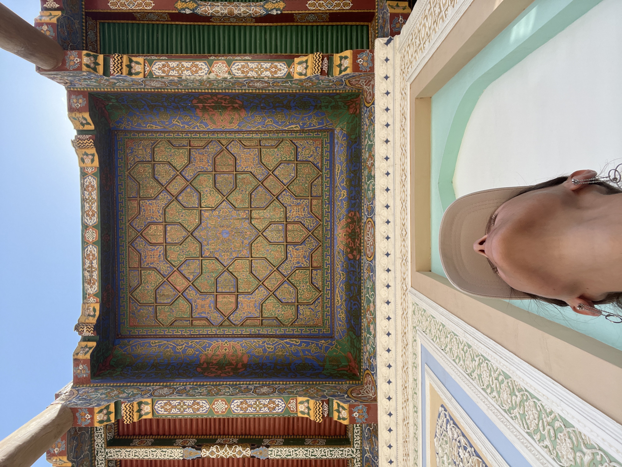 In Samarkand City, Uzbekistan, a person wearing a beige baseball cap and dangly earrings looks up at a highly ornate, colorful, and intricately designed ceiling. The ceiling features geometric patterns, floral motifs, and a rich palette of blues, greens, reds, and yellows. Below the ceiling, a white decorative border with blue diamond shapes is visible, leading to a beige architectural element with a light green arched opening. The perspective is from below, emphasizing the scale and detail of the architecture. The sky is visible above the structure.