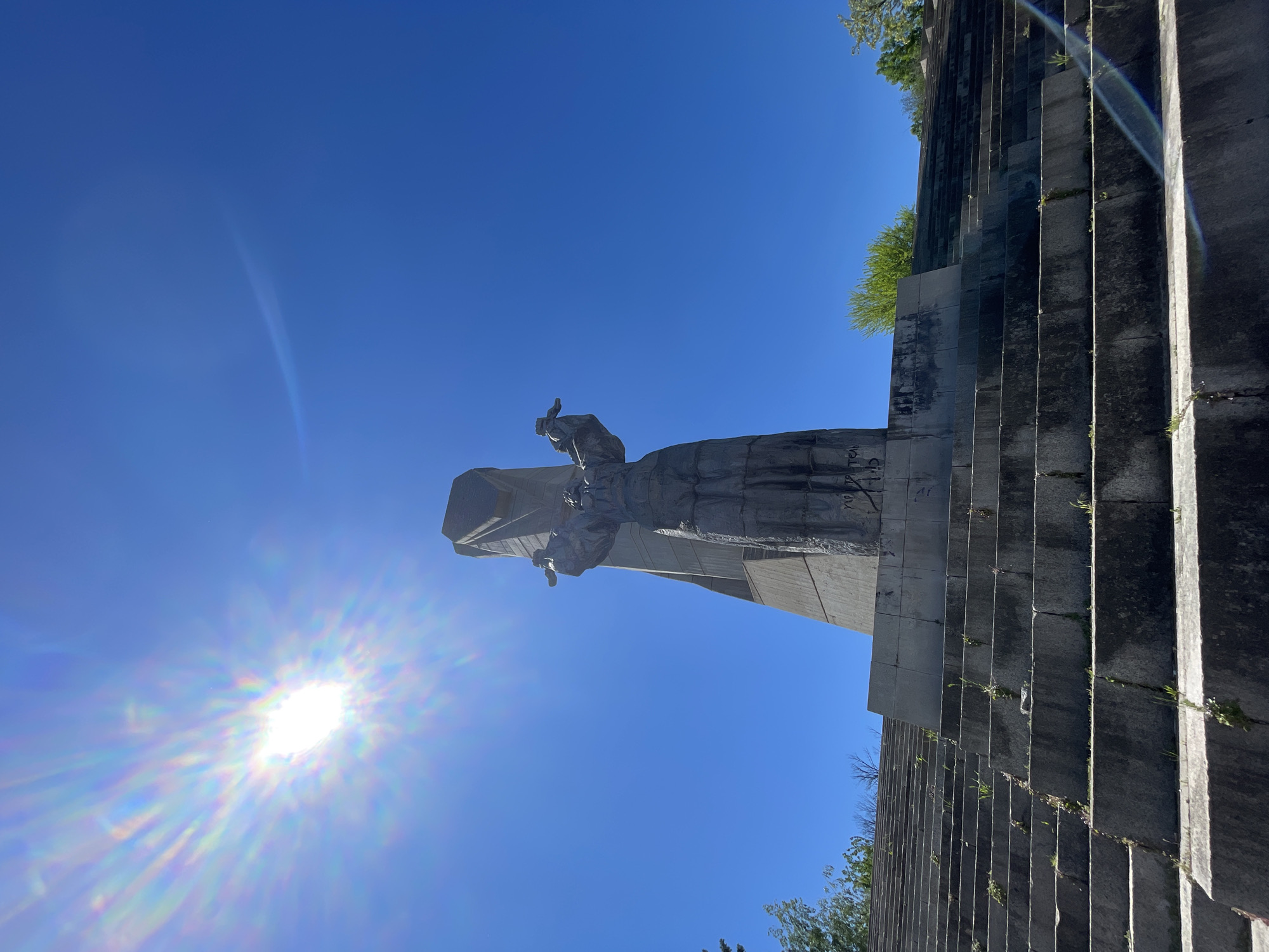 A low-angle shot depicts a gray statue against a bright blue sky with the sun visible in the lower left corner. The statue, situated to the left of a massive staircase, is composed of multiple figures, one of which appears to be raising its arm. The staircase is made of weathered stone. Green vegetation is sprouting in various places. The photograph is taken on a sunny day. The statue is located in Unknown, Unknown.