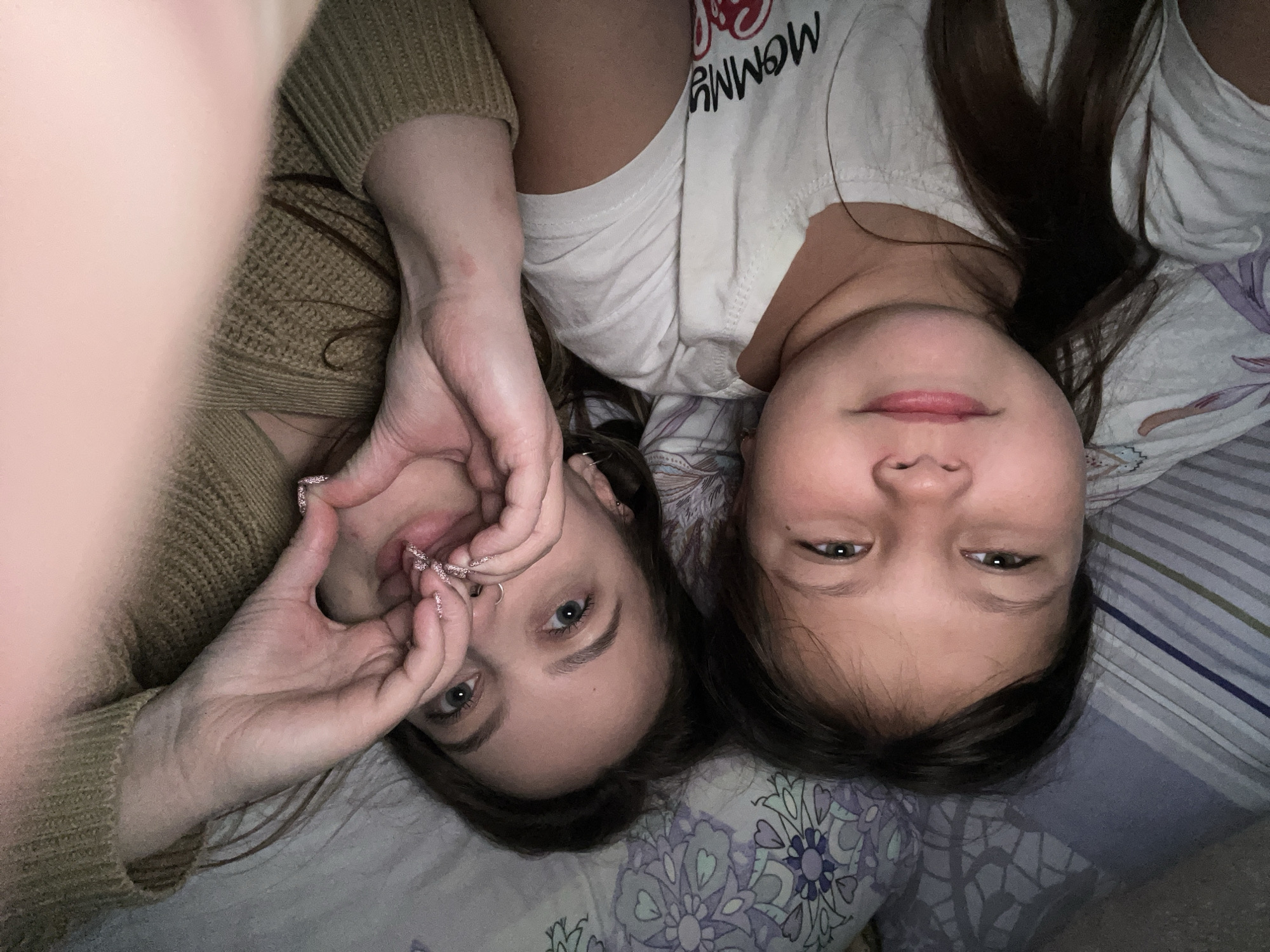 Two individuals, an adult female and a younger female child, are lying on a patterned bedding surface. The adult female is wearing a brown sweater and has her hands positioned to form a heart shape near her face. The younger female is wearing a white t-shirt with text. The location is Novopokrovskaya, Russia. The bedding has striped and floral patterns.