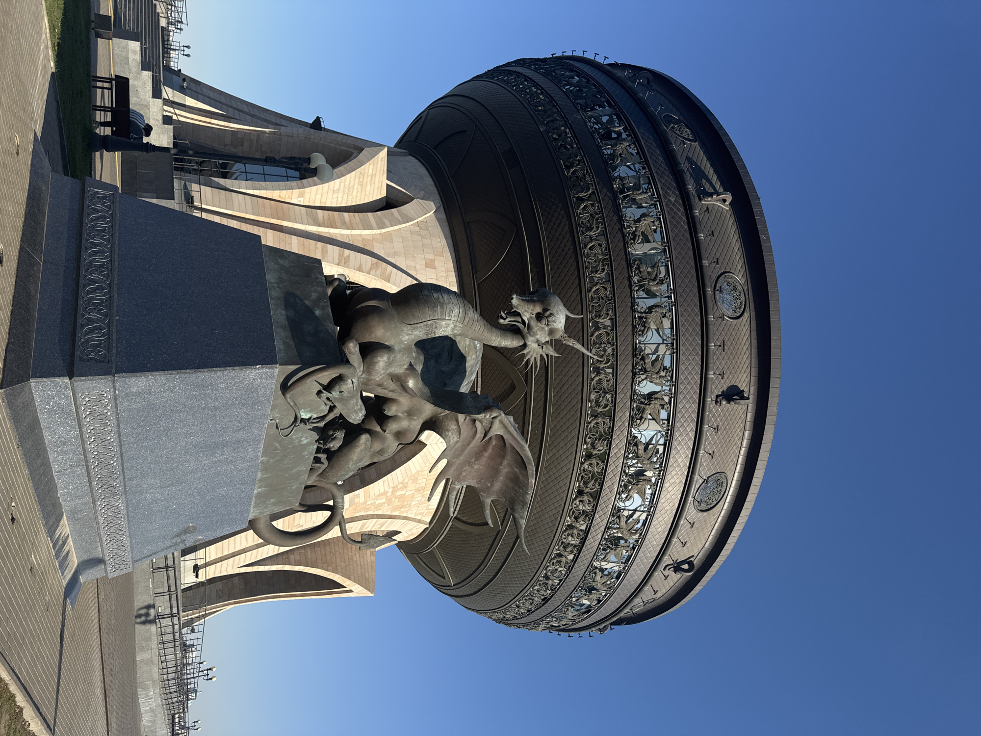 The image shows a statue of a dragon in Kazan, Russia, situated in front of a modern building with arched elements and a large, circular, dome-like structure adorned with intricate patterns and dragon motifs. The sky is clear and blue. A person is partially visible near the base of the building on the left side of the frame. The statue appears to be made of metal.