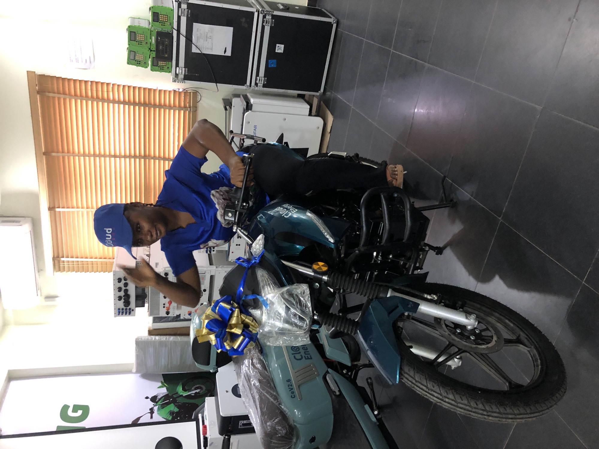 A woman wearing a blue shirt and a blue cap sits on a teal motorcycle in a showroom, likely in Ikeja, Nigeria. She is raising her right hand, gesturing towards the camera. The motorcycle is decorated with a large blue and gold bow. In the background, several pieces of equipment are visible, including large black cases, white battery units, and green electronic devices stacked on top of each other. A large white banner with green lettering and an image of a green motorcycle is mounted on the wall to the left. The floor is tiled with dark gray squares. The environment appears to be an indoor commercial space, possibly a showroom for electric vehicles or energy solutions.