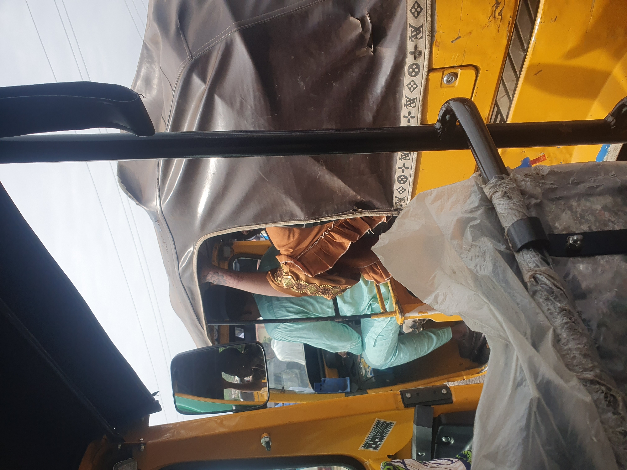 From the perspective of someone inside a yellow auto-rickshaw in Lokoja, Nigeria, a passenger wearing a brown and gold top with light blue pants is visible in another auto-rickshaw beside it. The view is framed by the rickshaw's black frame and brown canvas top, with a side mirror visible. Other rickshaws and occupants are visible through the window.
