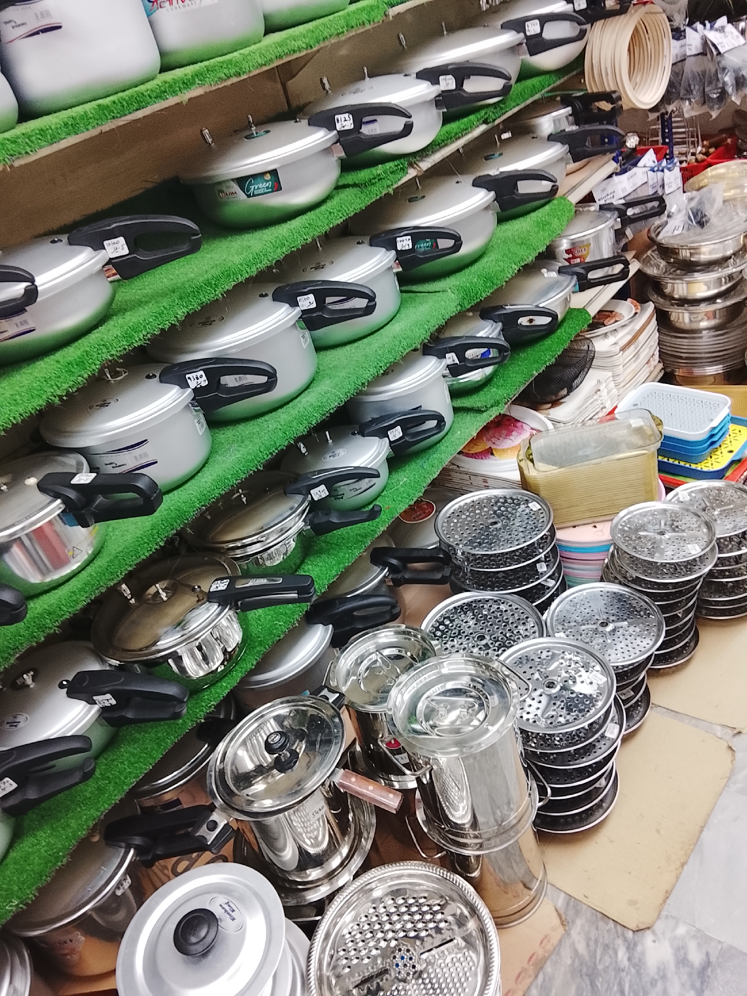 The image shows a retail display of kitchenware, likely in a shop in Gulgasht Colony, Pakistan. The shelves are lined with artificial green turf, and stocked with aluminum pressure cookers and other stainless steel kitchen tools such as graters and strainer sets. The pressure cookers, mostly silver, have black handles. Prices are affixed to some of the items. Other housewares, including plastic containers and metal bowls, can be seen in the background. The setting is a storefront.