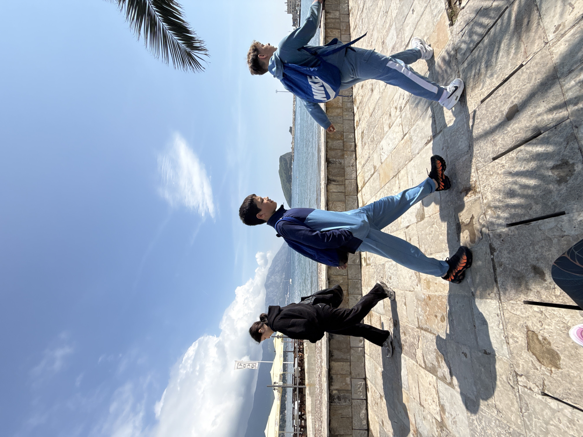 This image, captured in Budva, Montenegro, displays three individuals walking along a sunlit stone promenade next to the sea. The photo appears to be taken during a bright, sunny day, evidenced by the clear blue sky with scattered white clouds and the long shadows cast on the pavement.
In the foreground, on the left, a person in a black hoodie and black pants with dark hair tied back walks while looking slightly down or forward, wearing light-colored sneakers. Slightly ahead and to their right, a young male dressed in a blue and light-blue tracksuit looks upwards and towards his right, wearing black and orange sneakers. Further to the right, a second young male, also in a light blue tracksuit and carrying a blue backpack, mirrors the pose of the middle person by looking upwards and to his right. This individual wears white sneakers.
To the right of the promenade, a calm, blue sea stretches into the distance, with mountains visible across the water in the background. A dark green palm frond is partially visible in the upper right corner of the frame. On the left side of the promenade, an area with light-colored umbrellas and tables suggests an outdoor cafe or restaurant, and a white sign with illegible text can be seen in this vicinity. The word "NIKE" is clearly visible in white on the blue backpack of the person furthest to the right. The overall atmosphere is relaxed and leisurely.