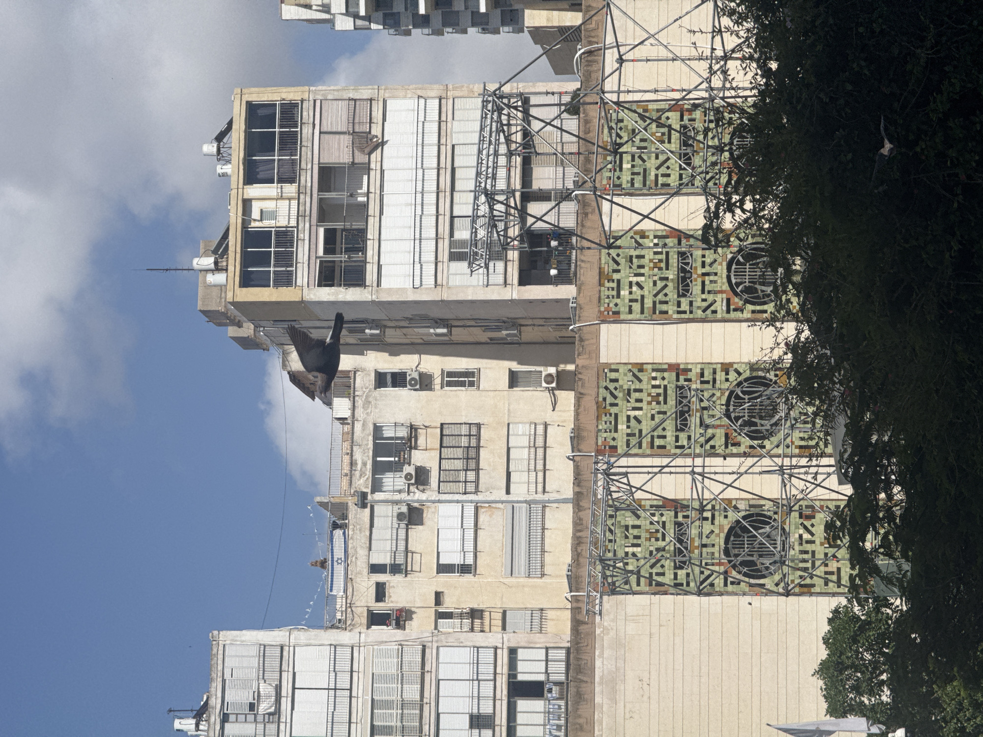 The image shows a multi-story apartment building in Ramat Gan, Israel. The facade has a mix of windows, some with shutters, and air conditioning units. A large tiled section with a geometric pattern and circular vents is visible, partially obscured by scaffolding and trees. An Israeli flag hangs from one of the balconies. A bird is captured mid-flight in front of the building. The sky is blue with scattered clouds. Other buildings can be seen in the distance.