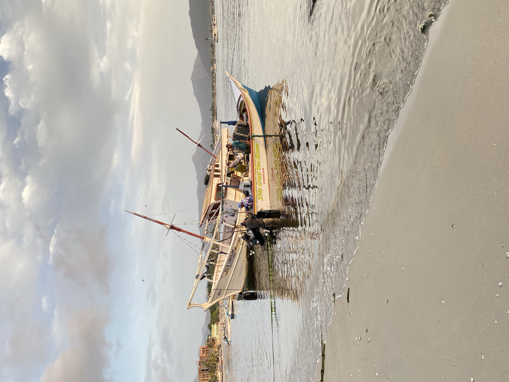 In Juan Griego, Venezuela, two fishing boats are anchored near a sandy beach. The larger boat, "JULIA CAROLINA," is white with blue trim and has "2850-FL 0312" written on its hull. A smaller boat, yellow with blue trim, is partially submerged and tied to the larger vessel. Both boats have people on board. The sky is filled with dramatic clouds, and mountains are visible in the distance. The water is calm, reflecting the sky and boats. The beach is sandy and scattered with shells.