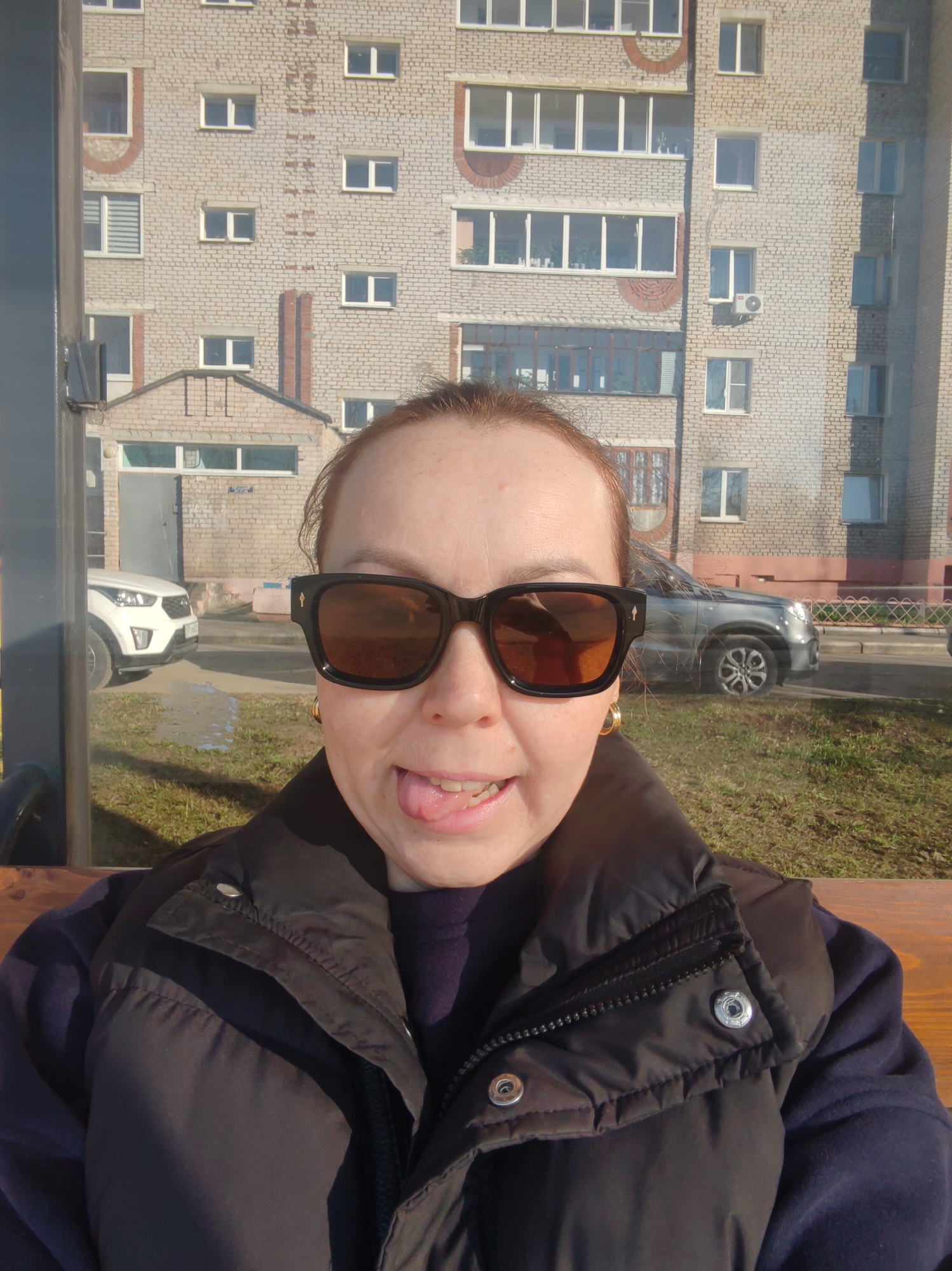 A woman is taking a selfie in front of a building in Chaikovsky, Russia. The woman is wearing sunglasses, a black vest over a blue sweater, and gold hoop earrings. She is sticking out her tongue. The background features a multi-story brick building with multiple windows, a white car, and a gray car. The setting appears to be a bus stop or waiting area, with a bench visible in the foreground. It is daytime, and the lighting suggests a sunny day. There is green grass and some brown patches of ground visible.
