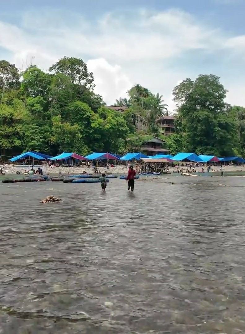 A wide shot captures a bustling scene along a shallow, rocky riverbank in a tropical environment. Numerous blue and red tarps are erected, suggesting a temporary gathering or market. People are visible under and around the tarps, and several blue inflatable rafts are lined up on the bank. Two figures, one in a red shirt and the other in a green shirt, are wading in the river in the foreground. The background is densely forested with lush green trees, and several buildings are nestled among the trees on a hillside. The sky is partly cloudy. The scene conveys a sense of community activity and outdoor recreation in a natural setting.