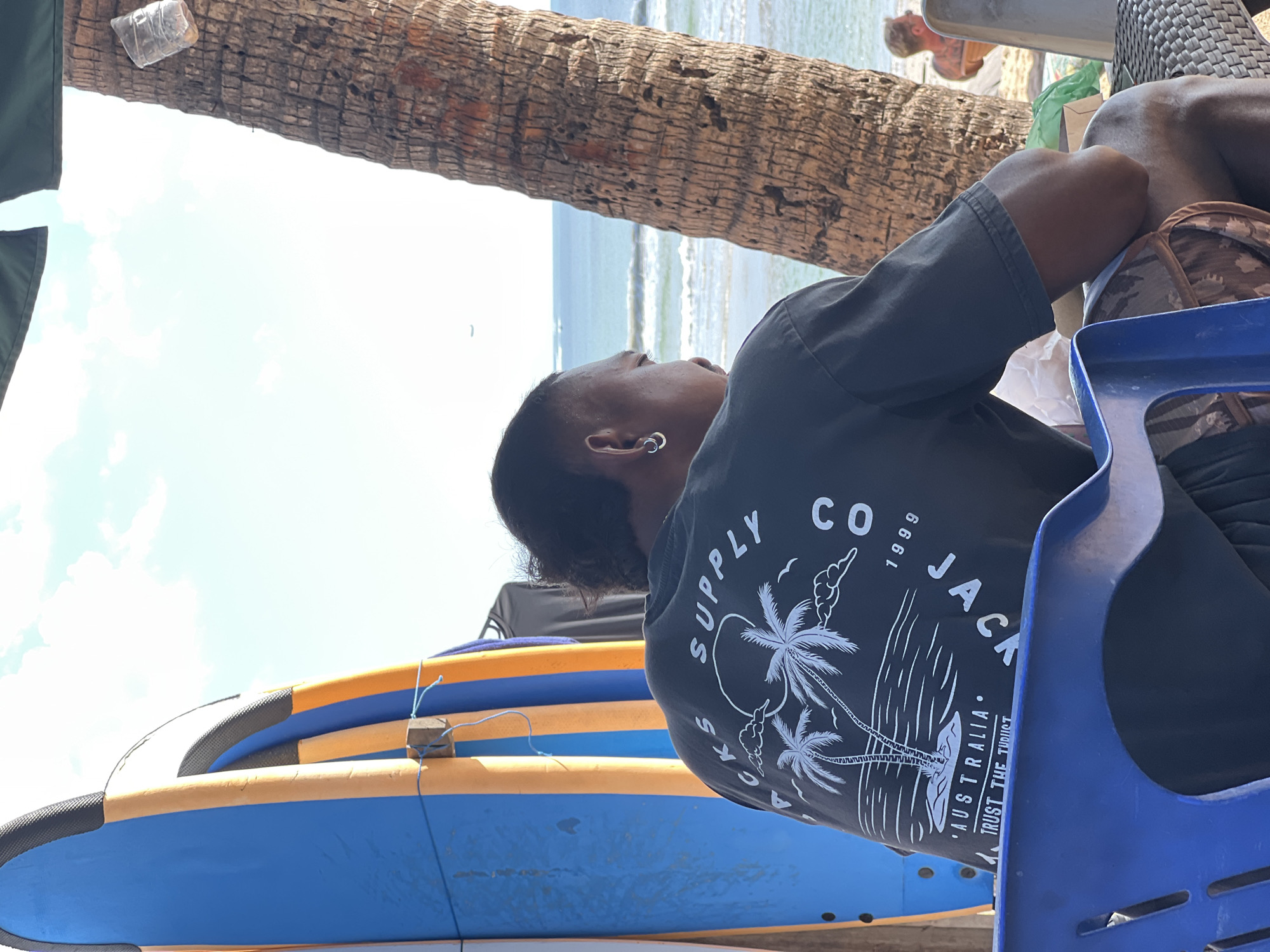 In Kuta, Indonesia, a man sits on a blue plastic chair facing away from the camera, with surfboards leaning against a wall to his left. He wears a dark t-shirt with a graphic design featuring palm trees and text that includes "AUSTRALIA" and "TRUST THE TUBUGT". To his right, a palm tree trunk is visible, and beyond that, the ocean with waves and a faint airplane in the sky. The background suggests a beach setting with a person in the distance. The lighting indicates it is daytime.