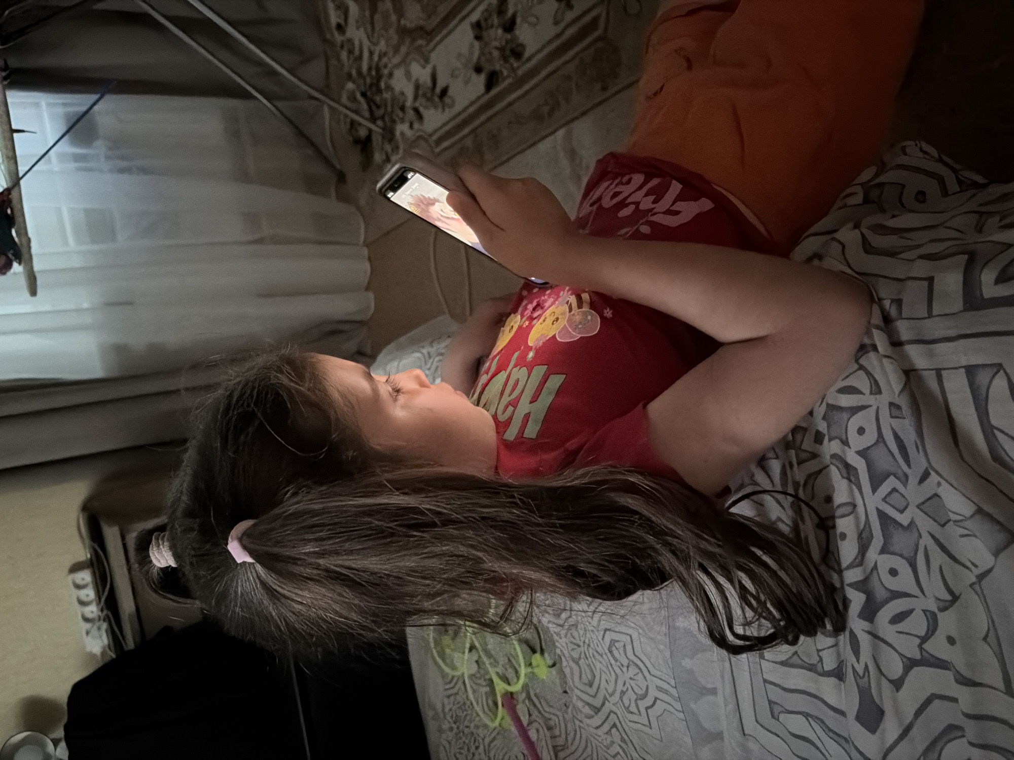 A young girl with dark hair tied in two ponytails lies on a bed, looking at a smartphone. She is wearing a red t-shirt with cartoon characters and orange shorts. The bed has a patterned grey and white sheet. In the background, a power strip with multiple outlets is visible on a shelf, along with a black fabric item. A curtained window and a patterned rug are also present. The scene appears to be indoors, likely a bedroom. The lighting suggests it is evening or night. No specific location in Tambov, Russia is identifiable from the image.