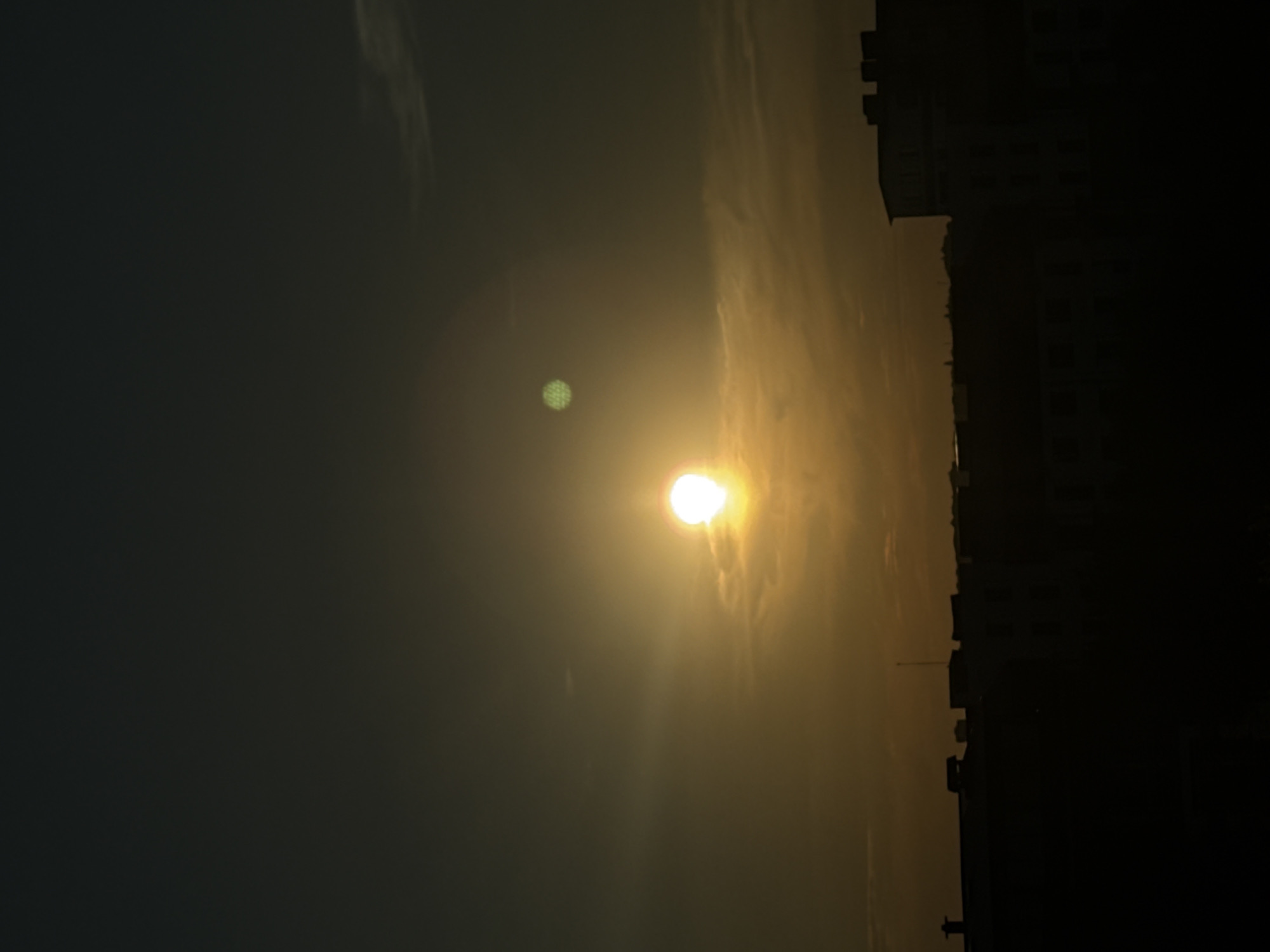 The image depicts a sunset or sunrise over a silhouetted cityscape. The sun, a bright, glowing orb with visible rays, is positioned centrally in the frame, casting a warm, golden light across the sky. There is a lens flare in the shape of a honeycomb pattern above and to the left of the sun. The sky transitions from a dark, hazy gray on the left to a bright, yellowish hue near the horizon. Wispy clouds are faintly visible, adding texture to the sky.
On the right side of the image, the silhouettes of buildings rise against the bright sky, creating a stark contrast. The buildings appear to be multi-story structures, possibly apartments or offices, with visible windows and rooflines. The lower portion of the image is dominated by the dark silhouette of the cityscape, with some structures appearing taller than others. The overall scene suggests an urban environment.
The lighting indicates it is either early morning or late evening, given the sun's position and the atmospheric glow. The haziness in the sky might suggest fog, pollution, or a dust-laden atmosphere. The image conveys a sense of tranquility and the vastness of the sky meeting the man-made structures of a city. No people are visible in the frame. There is no visible text in the image.