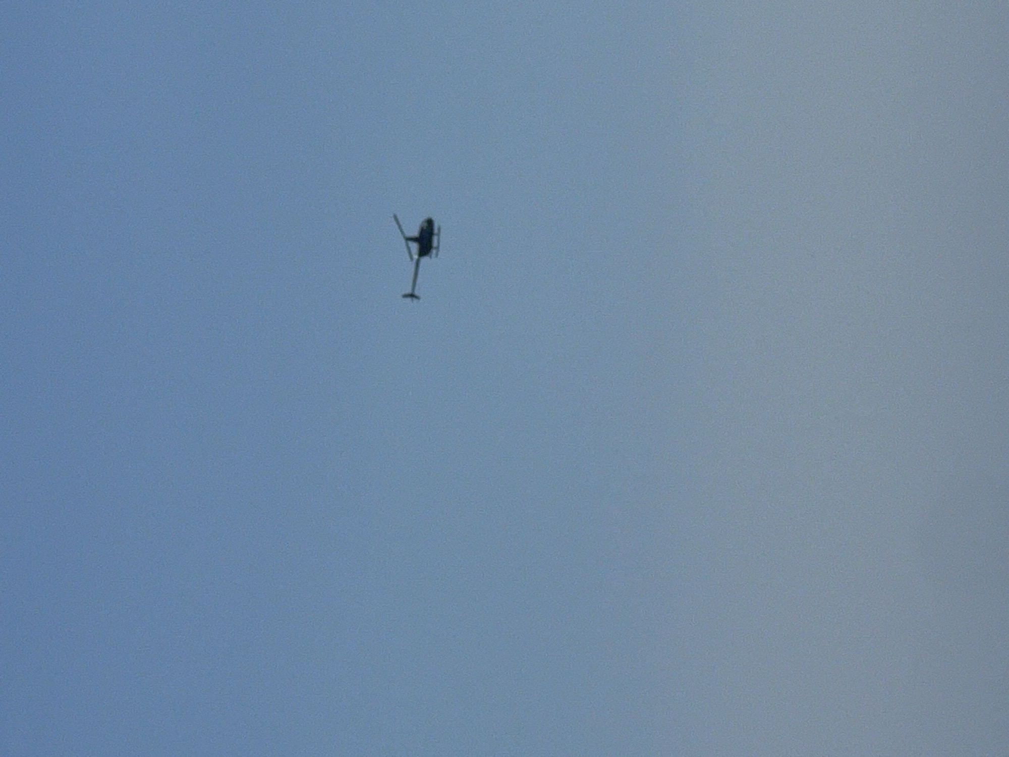 A single, dark helicopter is depicted in mid-flight against a clear, light blue sky. The helicopter is silhouetted and appears to be a small, two-bladed rotorcraft. It is positioned in the upper right quadrant of the image, with its tail rotor visible. The background is an uninterrupted expanse of sky, suggesting an aerial view. There is no indication of the city of Kuta, Indonesia within the image.