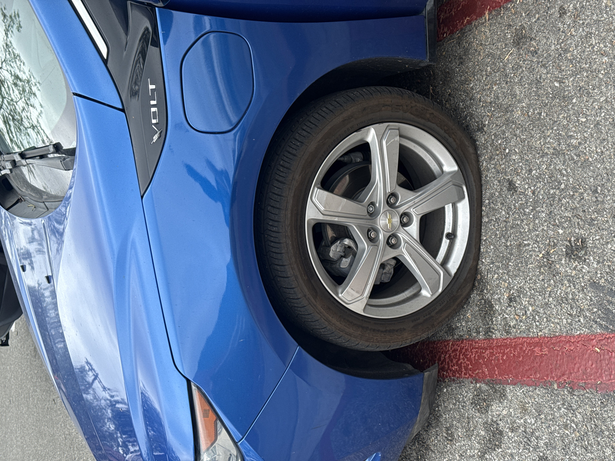 A close-up shot shows the front driver's side of a blue Chevrolet Volt, with the car parked partially over a red line painted on a gray asphalt surface. The word "Volt" is visible in silver lettering on the black trim of the car's fender. The car's tire is visible, along with a silver rim featuring the Chevrolet logo in the center. The tire tread appears worn. The vehicle is parked on what appears to be a street, with asphalt visible around the tire. The background is out of focus, making it difficult to discern the specific location within Los Angeles.