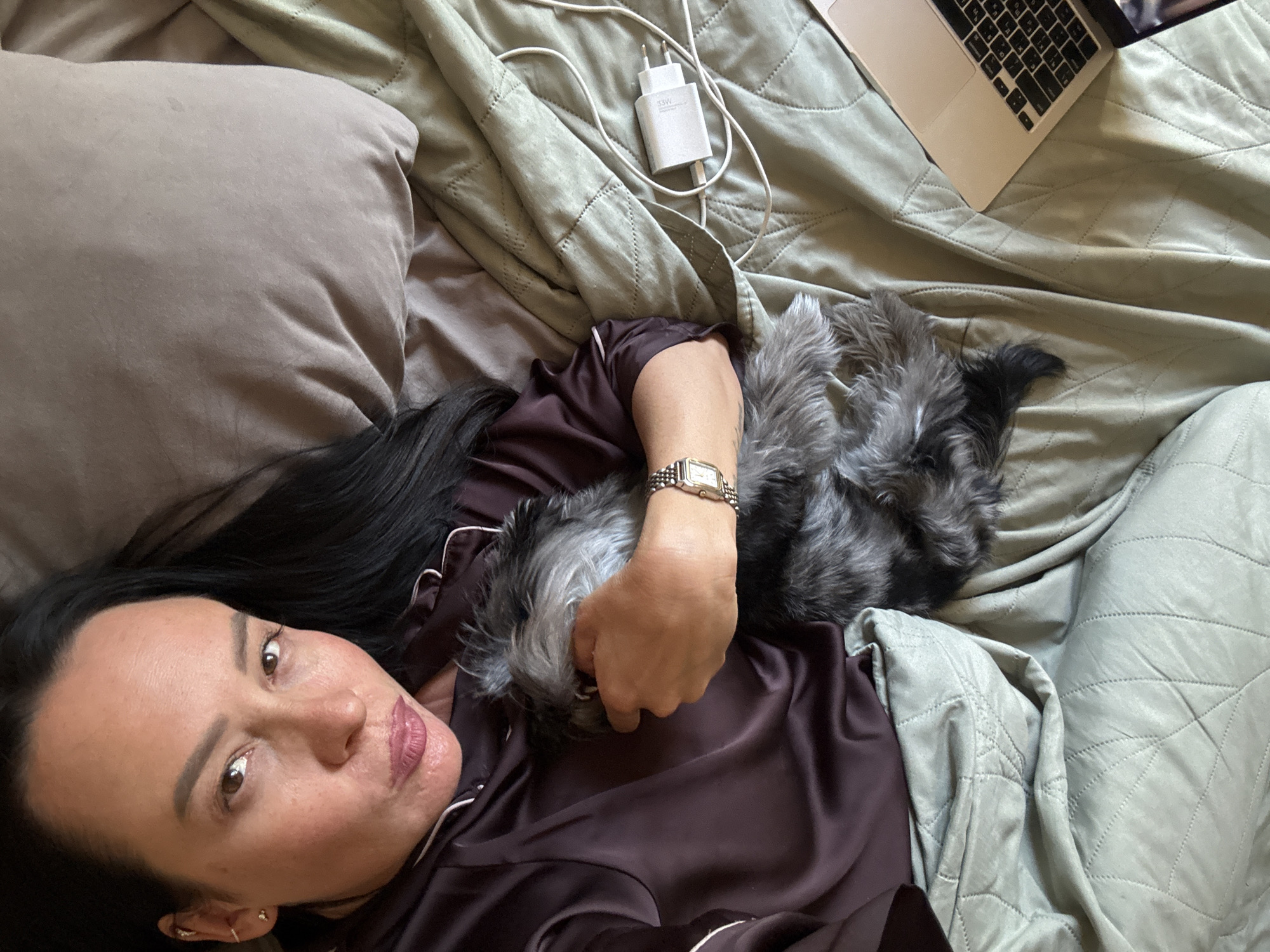 A woman lies on a bed, cradling a gray and black dog. She is wearing a brown pajama set with white piping and a silver watch on her left wrist. Her dark hair is spread on a pillow. Next to her on the bed is a laptop, a white USB charger, and a green quilted blanket. It is impossible to verify the location in Chaikovsky, Russia from this image alone.
