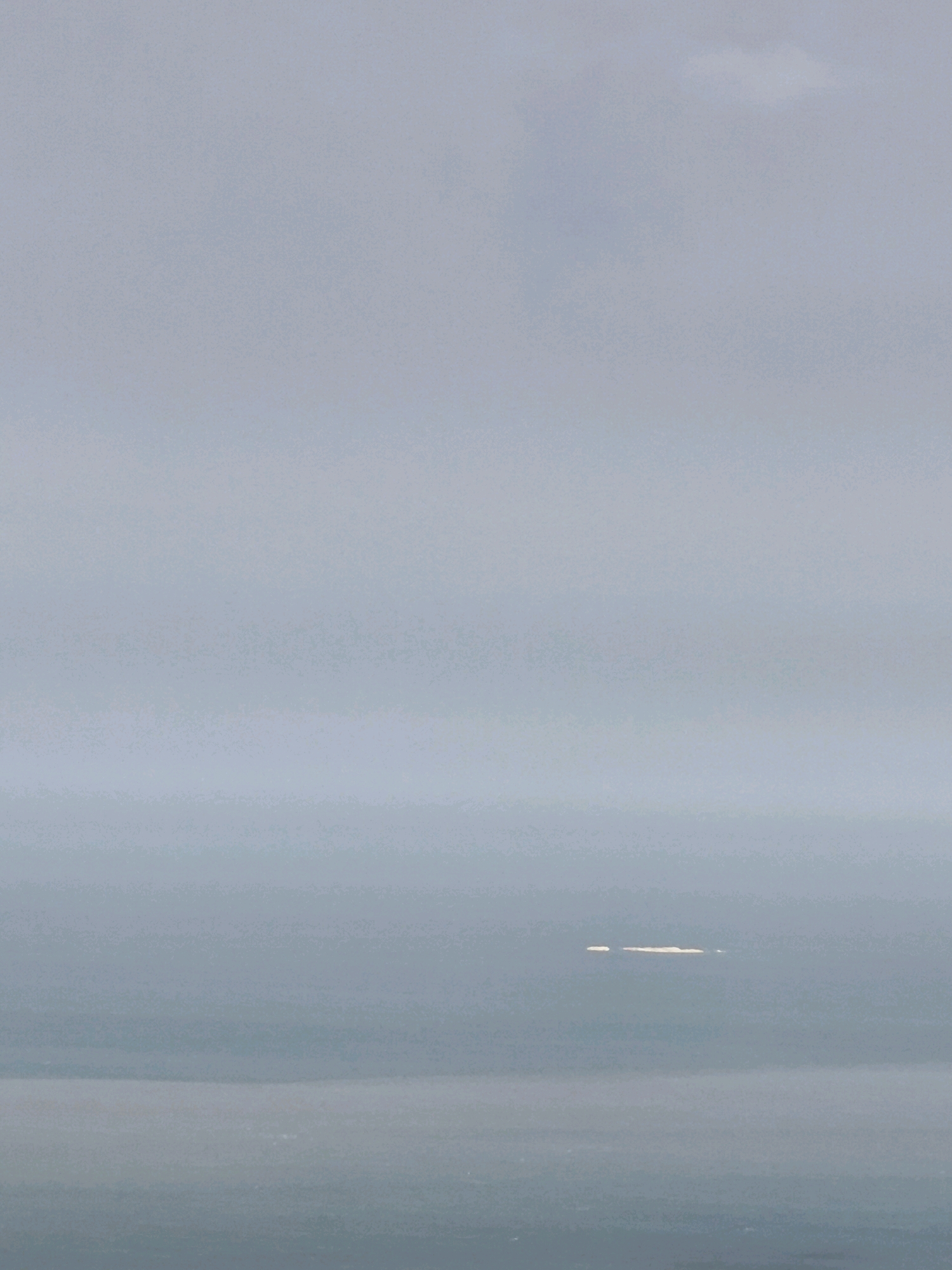 An aerial, wide shot captures a hazy, grey sky above a calm, expansive body of water. In the distance, a low-lying island, appearing as a light-colored, elongated shape, rests on the horizon. The water's surface is largely unbroken, with faint, darker horizontal lines suggesting gentle swells or distant shorelines. The overall atmosphere is serene and muted, dominated by soft, cool tones. The location is the sea near Qalaşıxı, Azerbaijan.