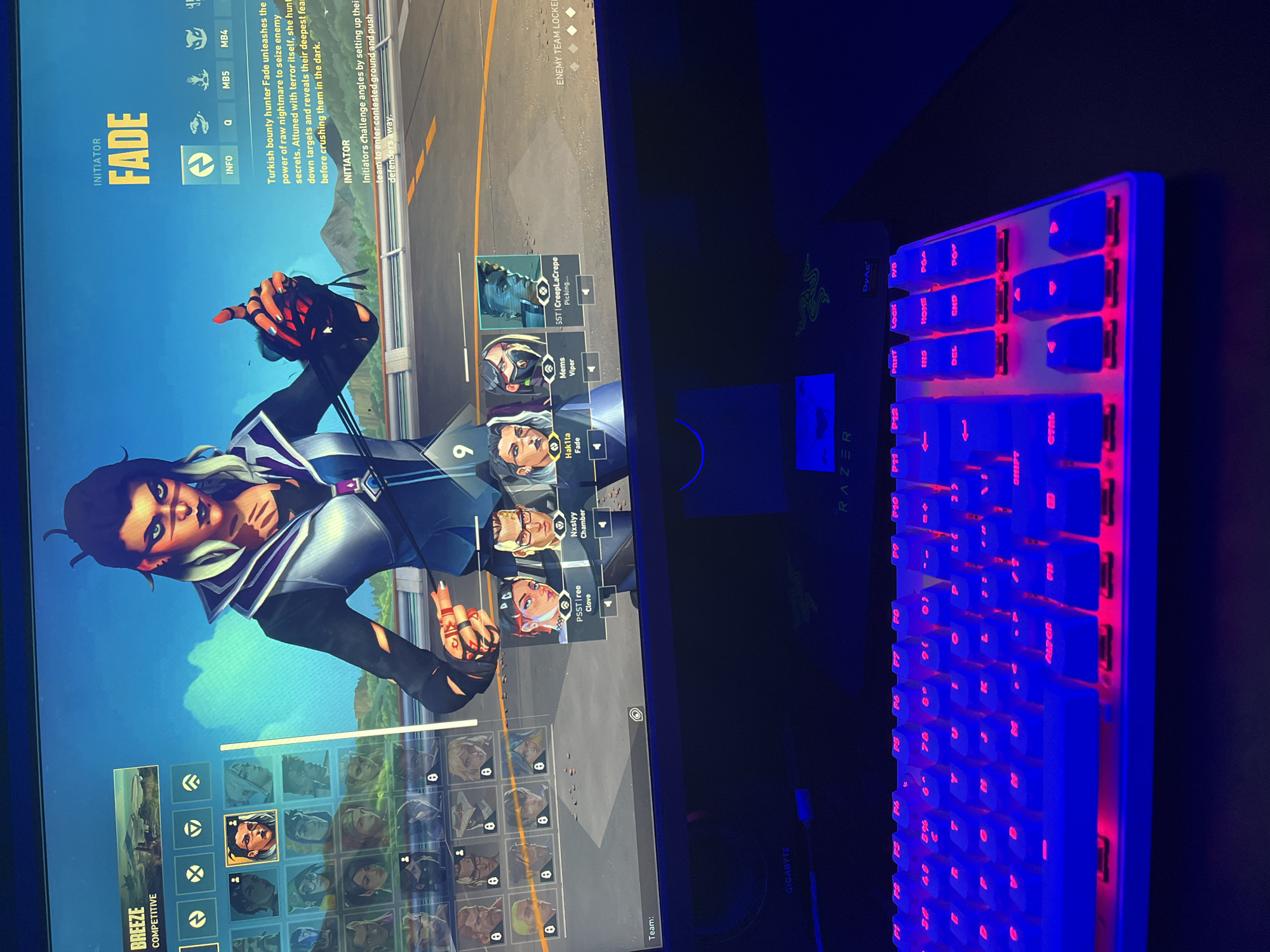 The image shows a person's gaming setup. A monitor displays the character selection screen of the video game Valorant, specifically the character "Fade." A Razer keyboard with red and blue backlighting sits to the right of the monitor. A Razer mousepad is visible under the keyboard. The environment is dark, with the light from the monitor and keyboard illuminating the desk area. No people are physically present in the frame. The screen displays information about the map "Breeze", a Competitive mode selection and a user named "Hak1ta" has selected the agent "Fade." The image also depicts agents like "Viper", "Chamber" and "Clove" being selected by teammates. The bottom portion of the image appears to depict other potential agents to select. The image does not depict anything related to the city of Burgas, Bulgaria.