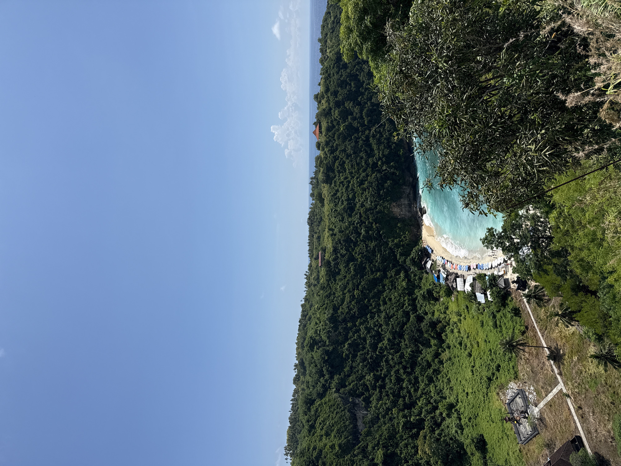 The image captures a coastal scene in Pejukutan, Indonesia. Lush, dense green vegetation covers the majority of the landscape, leading down to a beach with turquoise water. A small sandy beach is bordered by white-capped waves. Several structures, possibly huts or shacks, are situated near the shoreline, with a collection of colorful objects, likely beach umbrellas, arranged in a row along the beach. A path or walkway extends from the beach area toward an elevated point, where a structure or monument with dark-colored stone or concrete is situated. The sky is a clear blue with light clouds on the horizon. Trees and foliage are visible in the foreground, partially obscuring the view, suggesting the photograph was taken from a high vantage point, such as a cliff or elevated structure.
