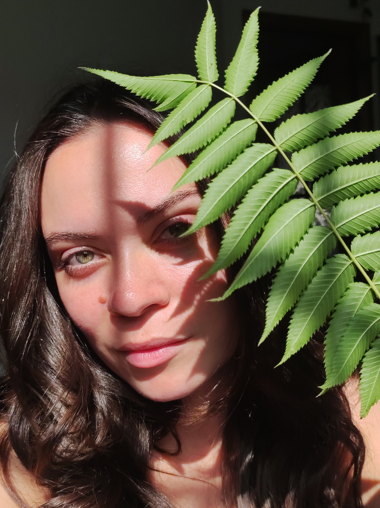 The image is a close-up portrait of a young woman with dark, wavy hair and green eyes. She is looking directly at the camera, with a branch of a green, serrated leaf plant obscuring the right side of her face and hair. Sunlight casts shadows across her forehead, nose, and cheek. Her skin appears fair and smooth, with a mole visible on her right cheek. Her lips are slightly parted and painted with a soft pink lipstick. The background is dark and out of focus, suggesting an indoor setting with natural light coming from the side. There are no visible landmarks that would indicate the location in Chaikovsky, Russia.
