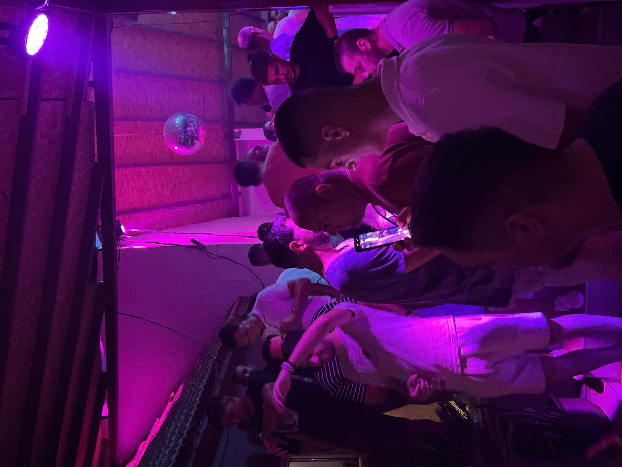 The image shows a group of people gathered in an indoor space illuminated by vibrant pink and purple lights. The environment appears to be a nightclub or bar, with a disco ball visible in the upper portion of the frame. Several individuals are standing and interacting, with some holding drinks. The clothing styles suggest a casual evening setting. The context provided indicates this scene is located in Herceg Novi, Montenegro.