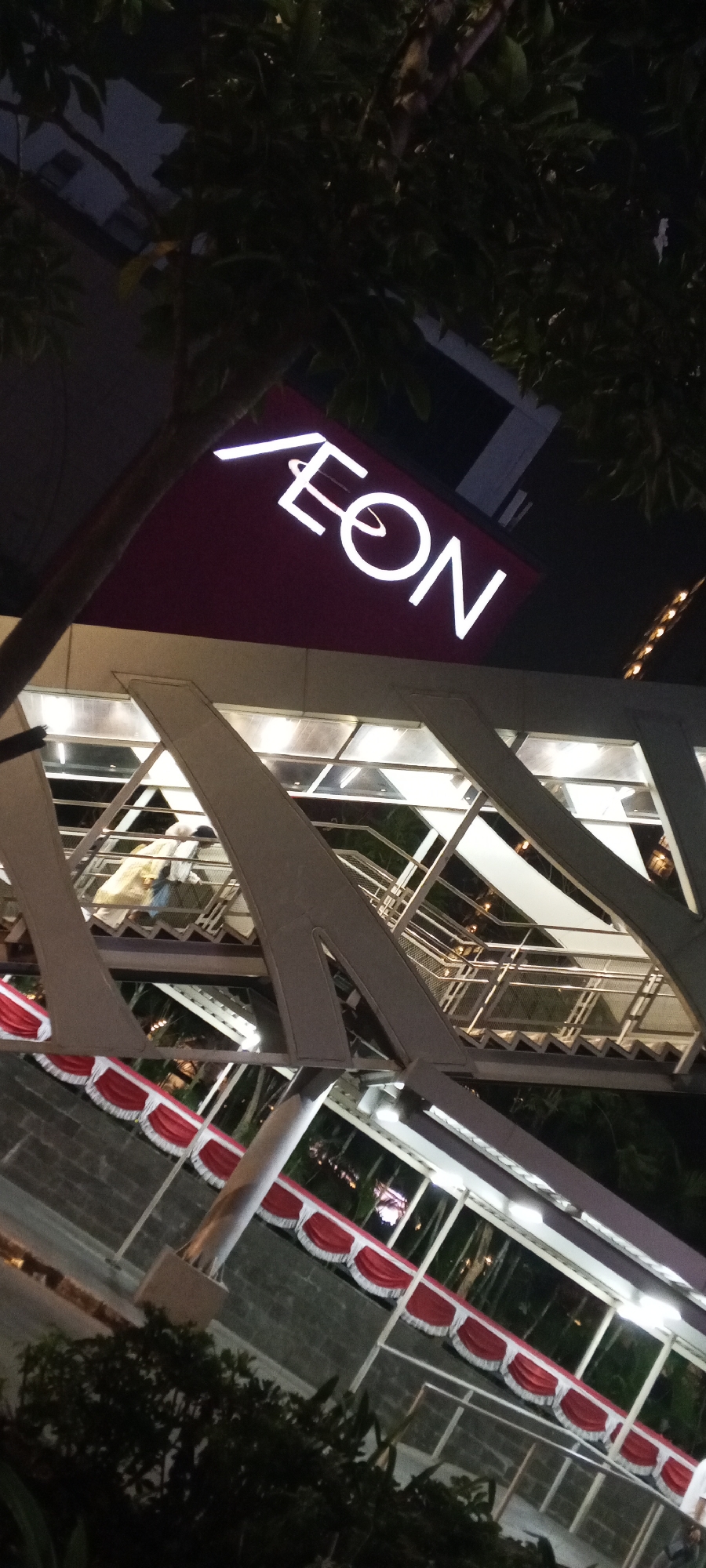The image shows the exterior of a building with a large, illuminated sign that reads "AEON". The sign is white neon against a dark red background. Below the sign is an elevated walkway or bridge with a modern, angular design. The walkway has metal supports and a transparent roof, allowing light from the interior to shine through. Several people are visible on the walkway, some appearing to be walking up stairs. The walkway is decorated with red and white banners, possibly for a national celebration. The scene is captured at night, with the surrounding environment appearing dark and dimly lit, with some trees visible in the foreground. The architecture and the presence of a large shopping mall sign suggest an urban commercial area. This is likely AEON Mall Tanjung Barat, located in South Jakarta, Indonesia, known for its distinctive architecture and as a prominent shopping destination.