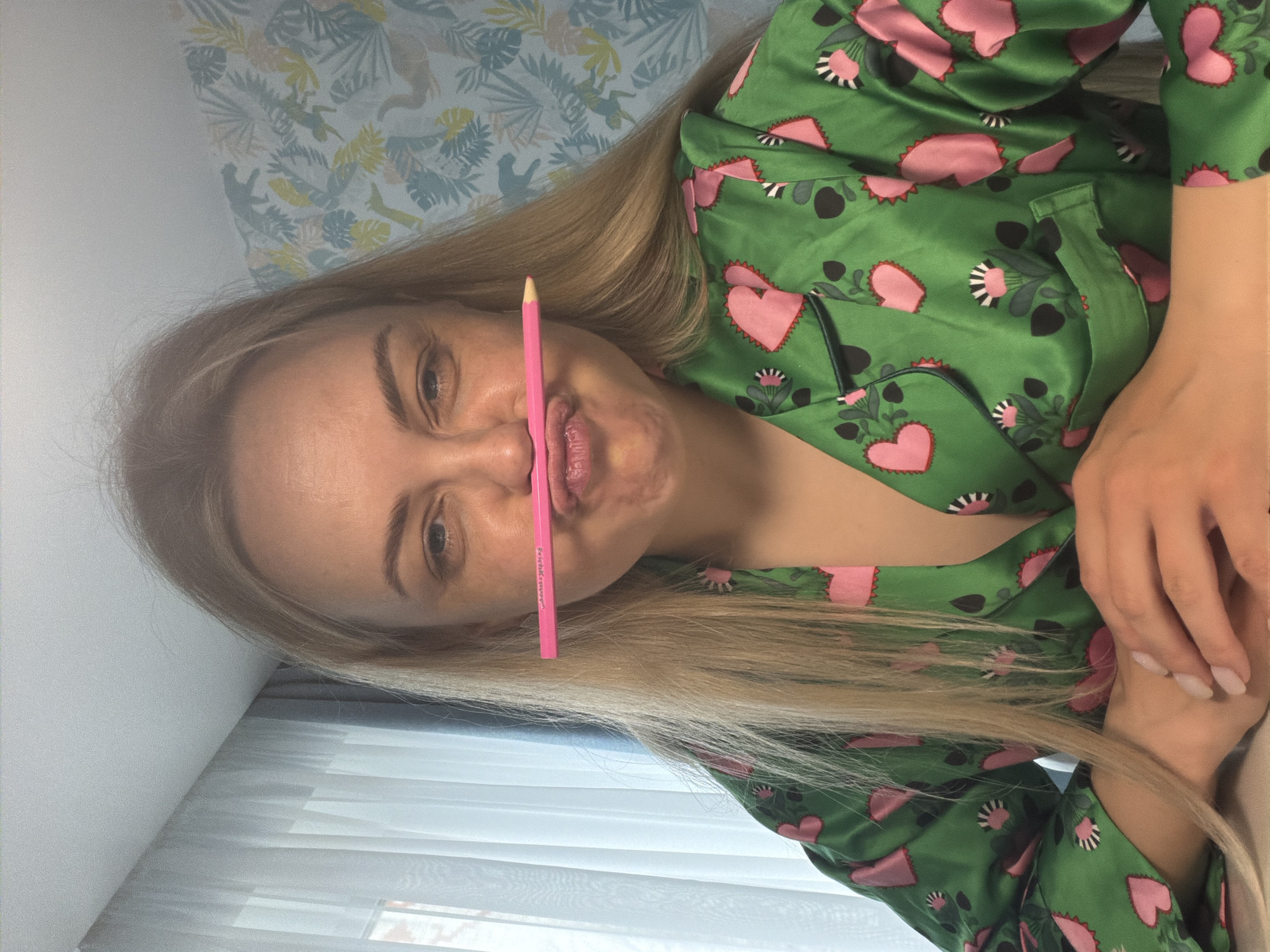 This is an indoor portrait of a young blonde woman from Votkinsk, Russia, captured with the camera rotated 90 degrees clockwise. The woman is looking directly at the viewer with a playful expression, holding a pink pencil horizontally between her upper lip and nose. Her lips are slightly puckered in a humorous manner.
She has long, straight blonde hair and is wearing a lustrous emerald green satin top, possibly pajamas, which is patterned with large pink hearts and smaller botanical elements in pink, black, and white. Her hands, featuring light-colored manicured nails, are visible and appear clasped together on her lap or chest.
The background shows an indoor setting. On the left side of the frame (when viewed upright), there is a wall with light blue and white wallpaper featuring a jungle-themed pattern of leaves and animal silhouettes, possibly monkeys or lemurs. On the right (when viewed upright), a sheer white curtain covers a window, allowing diffused light into the room. A plain white wall is also visible in the upper and lower portions of the frame. The pink pencil has faint text visible on its side, which reads "Essence Cosmetics".