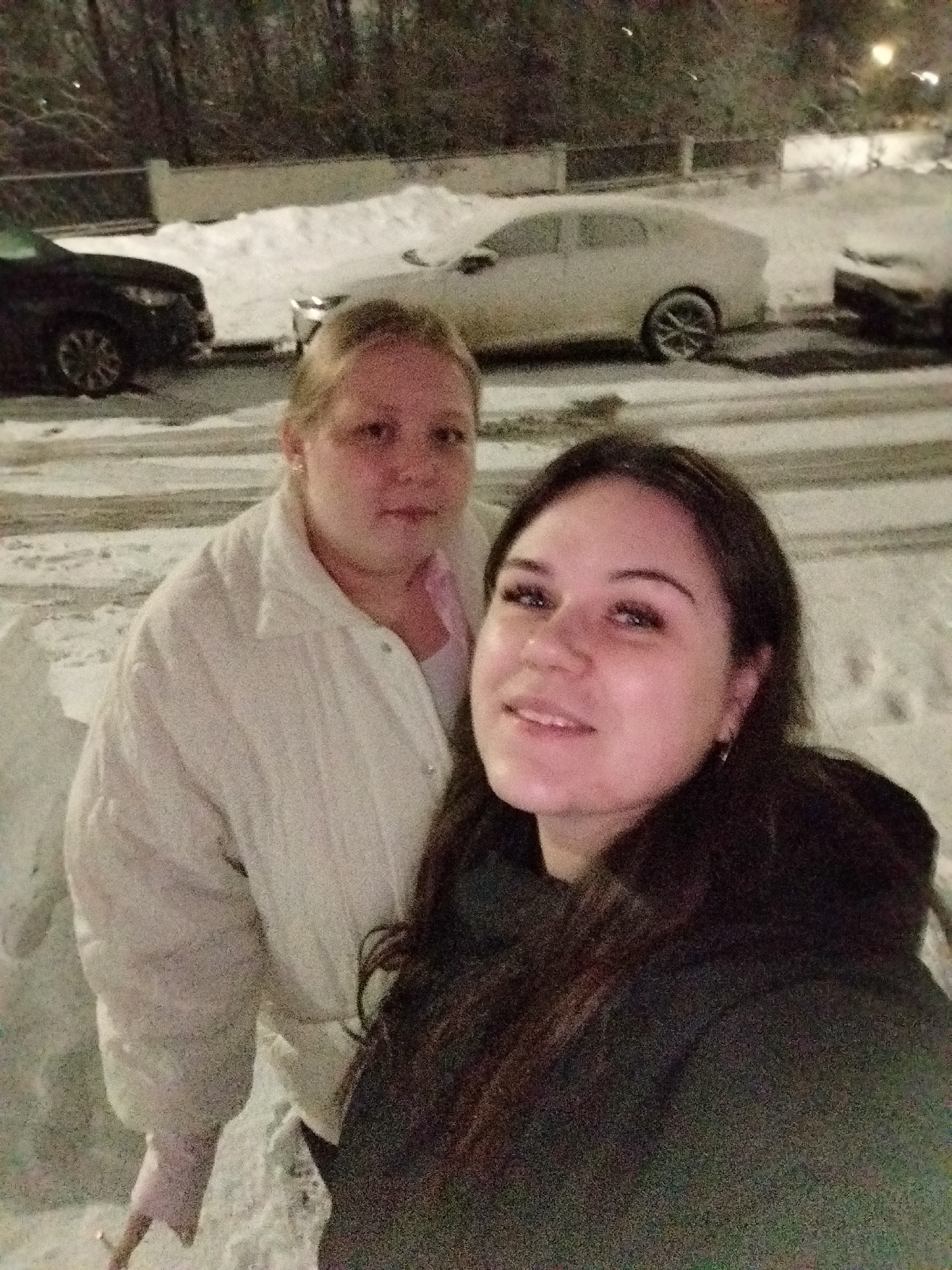 Two women are standing outside on a snowy street. The woman in the foreground has dark hair and is wearing a dark jacket. The woman behind her has blonde hair and is wearing a white jacket. There are parked cars covered in snow in the background, along with snow-covered ground and buildings. The photo was taken in Chaikovsky, Russia.