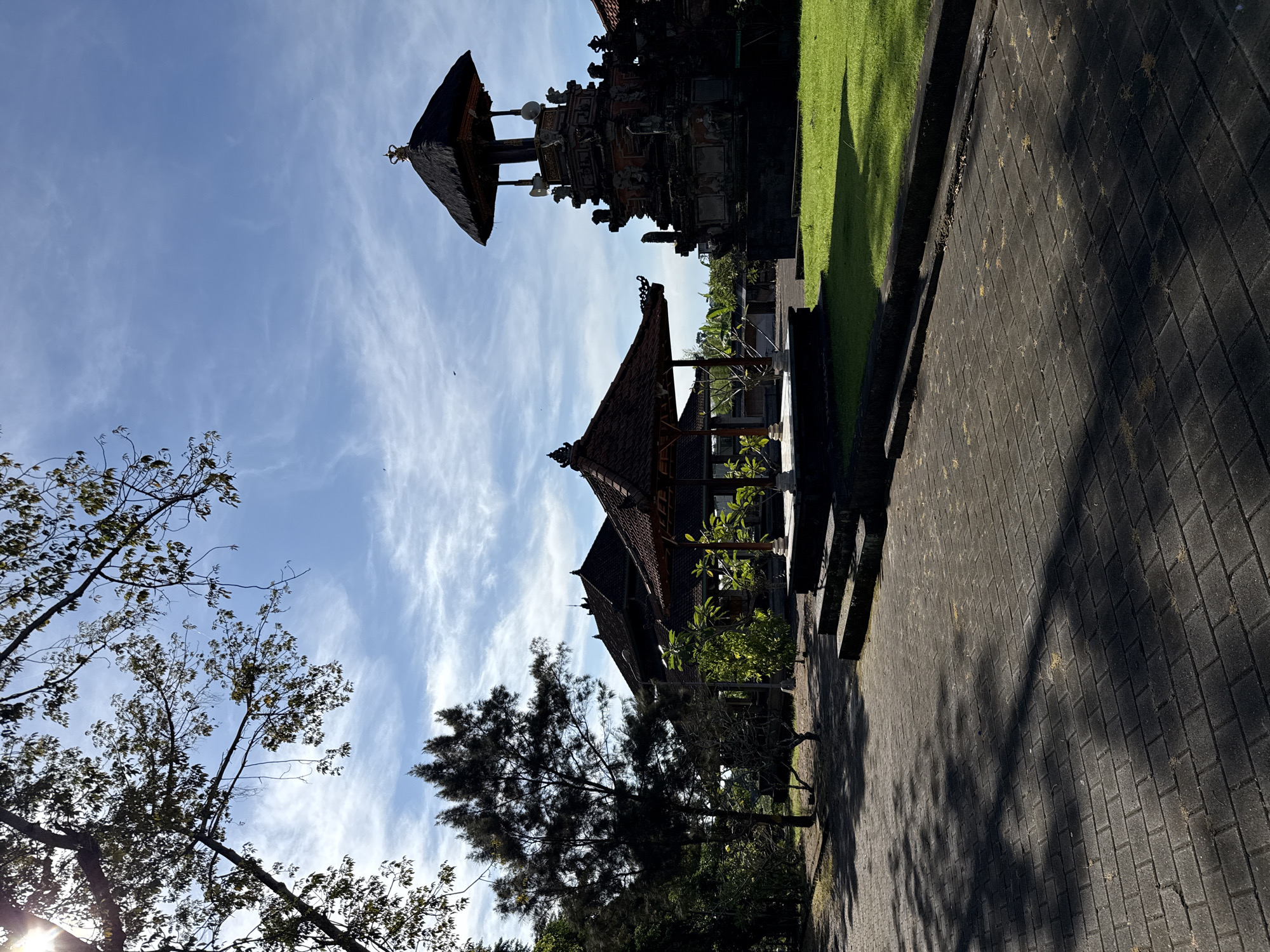 The image shows a scene in Mengwi, Indonesia. A paved pathway runs along the right side of the frame. The left side of the path is lined with grass and traditional Balinese architecture. The buildings are characterized by dark-tiled roofs and ornate stone carvings. Several trees are visible near the pathway. The sky above is blue with scattered white clouds.