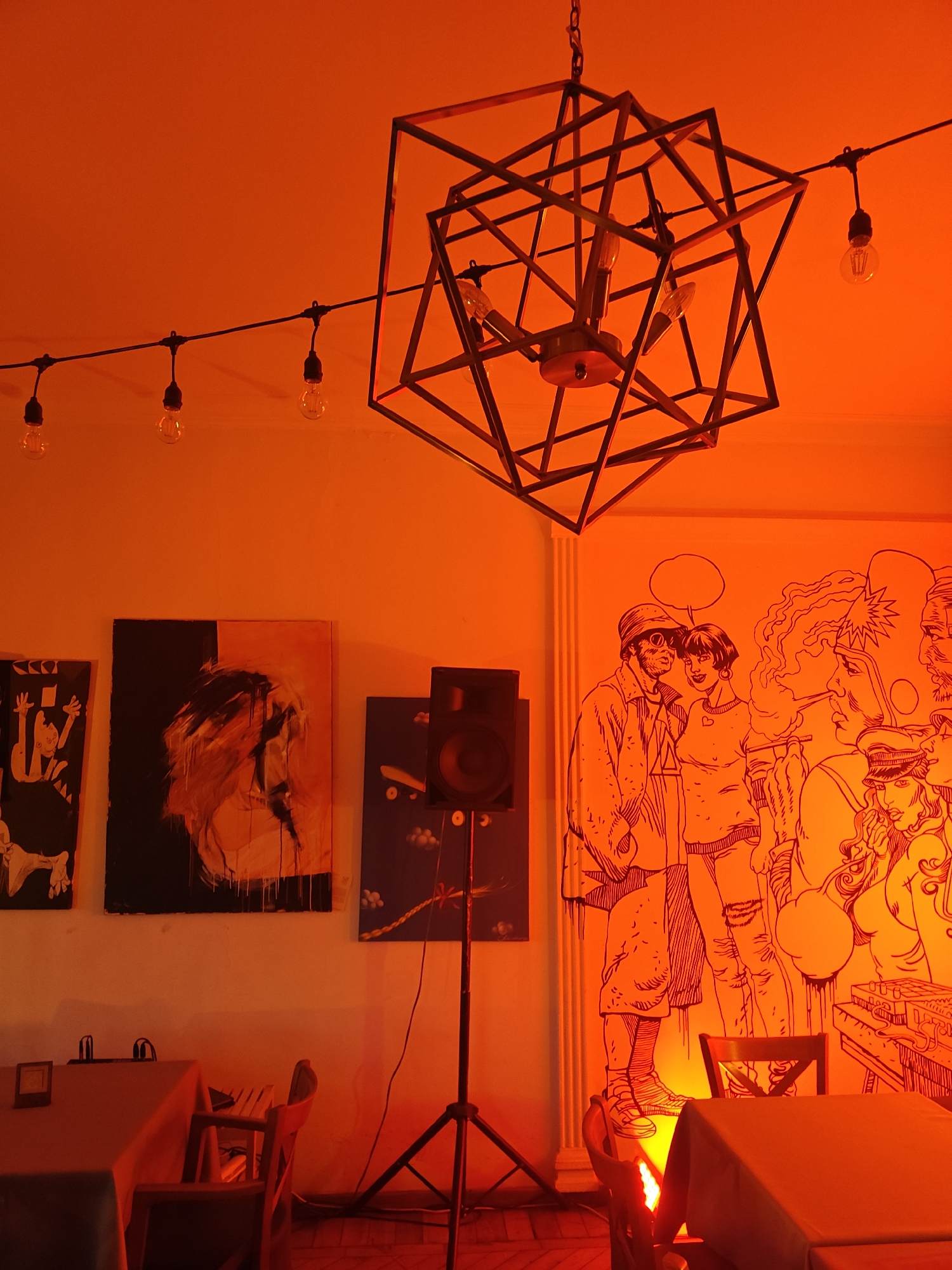 The image depicts an interior space illuminated with a strong red light. The location appears to be a restaurant or bar with art on the walls, including three framed paintings and a large mural depicting people. There is a geometric metal chandelier hanging from the ceiling, along with a string of light bulbs. A speaker is positioned on a stand in front of the artwork. The furniture visible includes tables and chairs, all covered in red light. There are no people explicitly shown, but the mural suggests human interaction as a central theme of the space. The city and region are unknown.
