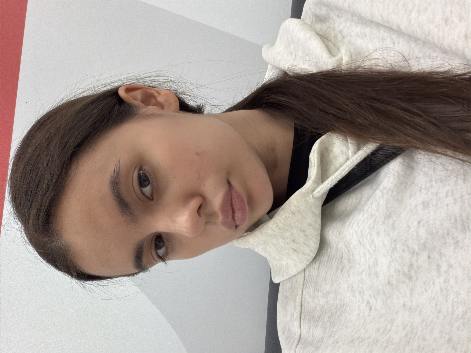 A close-up portrait shows a young woman with light skin, brown hair, and brown eyes. She is wearing a light-grey hooded sweatshirt with a black collar visible underneath. Her hair is styled straight and falls to her right side. The background is a plain white wall with a vertical red stripe visible in the top-left corner. There are no visible cues to determine the location in the city of Chaikovsky, Russia.
