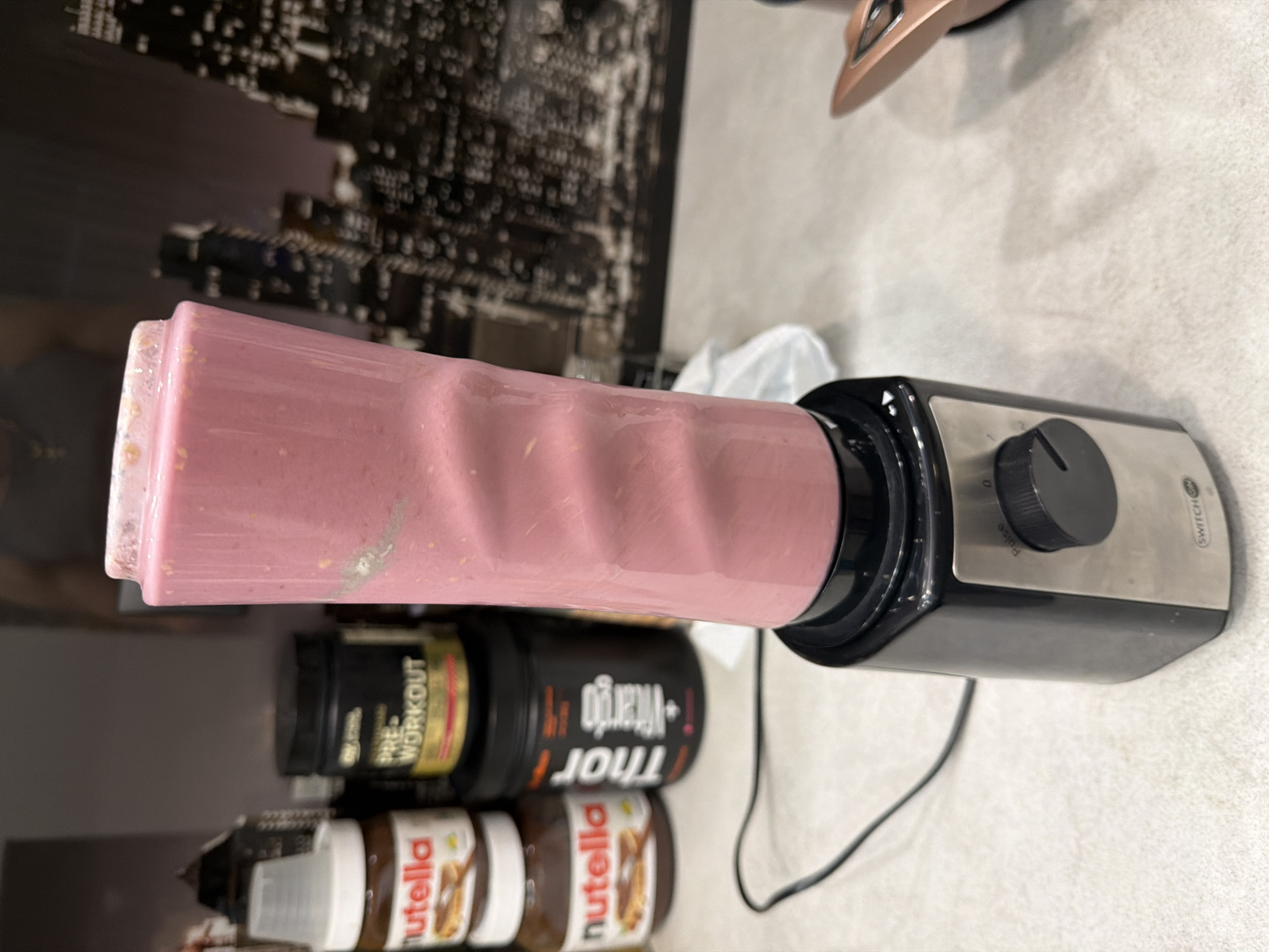 The image shows a blender with a pink smoothie in it, positioned on a surface near several containers of Nutella and pre-workout supplements. The background features an out-of-focus image, possibly a cityscape. There are no people visible. The exact location in Plovdiv, Bulgaria, cannot be determined from the image alone.