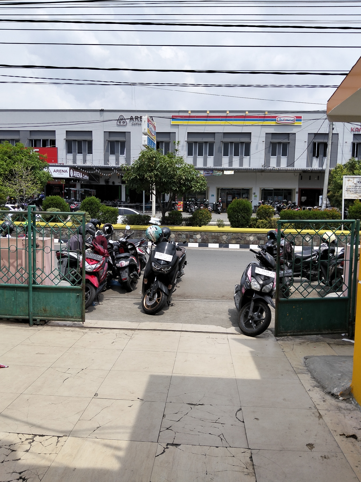 The image shows a view of a street in Bireuen, Indonesia, with a row of buildings in the background and several motorcycles parked in the foreground. The buildings appear to be commercial establishments, with signage indicating "ARENA Coffee" and "Indomaret." The street is paved, and there are power lines overhead. A green metal gate is partially visible on the left side of the image, and a yellow structure is on the right. The ground in the foreground is tiled and shows signs of wear and cracking. Several motorcycles are parked on the street and sidewalk area, some with helmets on them. The sky is overcast.