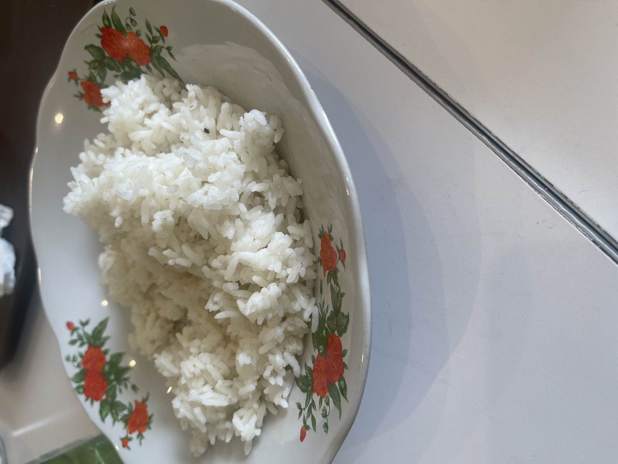 A white bowl with a floral pattern of orange and green flowers holds a mound of white rice. The bowl is positioned on a white surface, possibly a table, with a diagonal seam running through it. The lighting suggests an indoor setting, possibly a restaurant or home in Medan, Indonesia. No people or other objects are clearly visible.