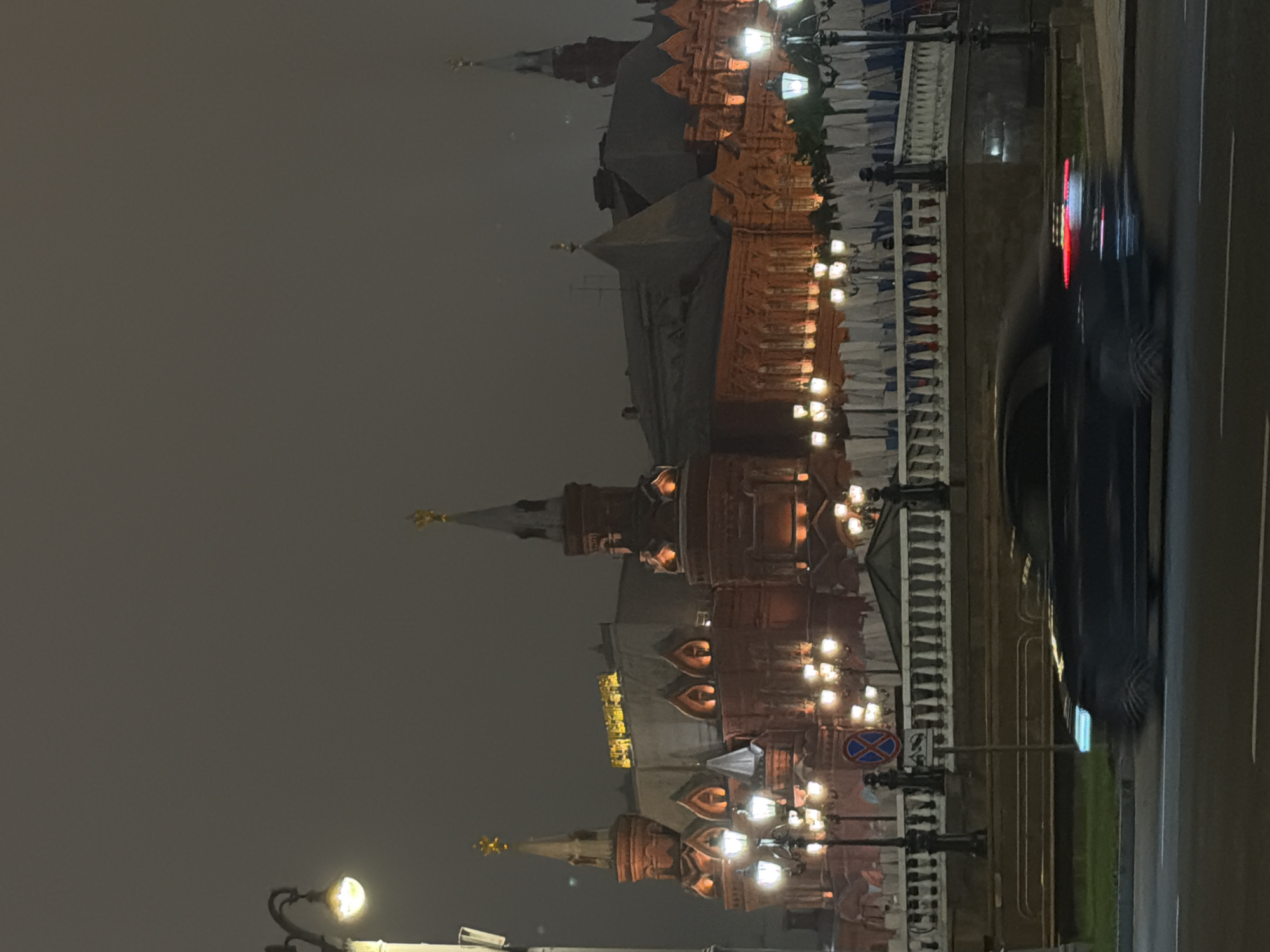 The image shows a night scene in Moscow, Russia, featuring the illuminated architecture of the Kremlin towers. A road runs along the right side of the frame, with a blurry car passing by. Streetlights are visible along a decorative fence bordering the road. The sky is overcast, providing a dark backdrop for the illuminated buildings.