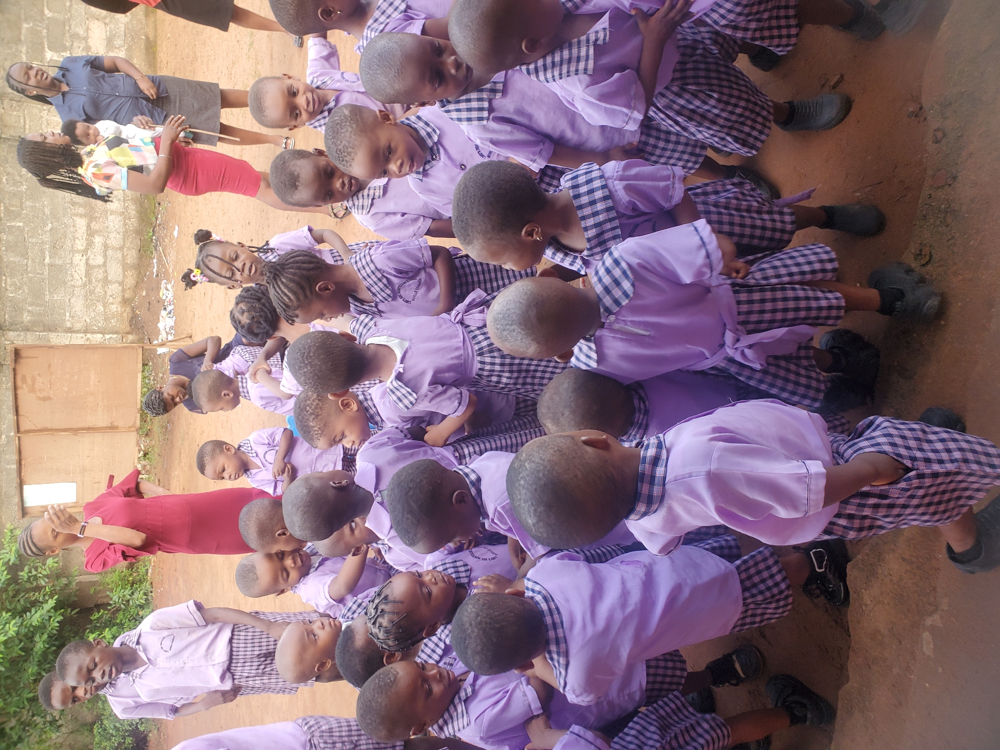 A group of school children dressed in lavender uniforms are seated on the ground in what appears to be an outdoor space in Ukwu-Abwa, Nigeria. The uniforms consist of a lavender top with checkered skirts or pants. A few adults are visible, including two women and a baby being held by one woman. One woman wears a blue top and gray skirt while the other wears a red dress. The surrounding environment includes a wall and some greenery.