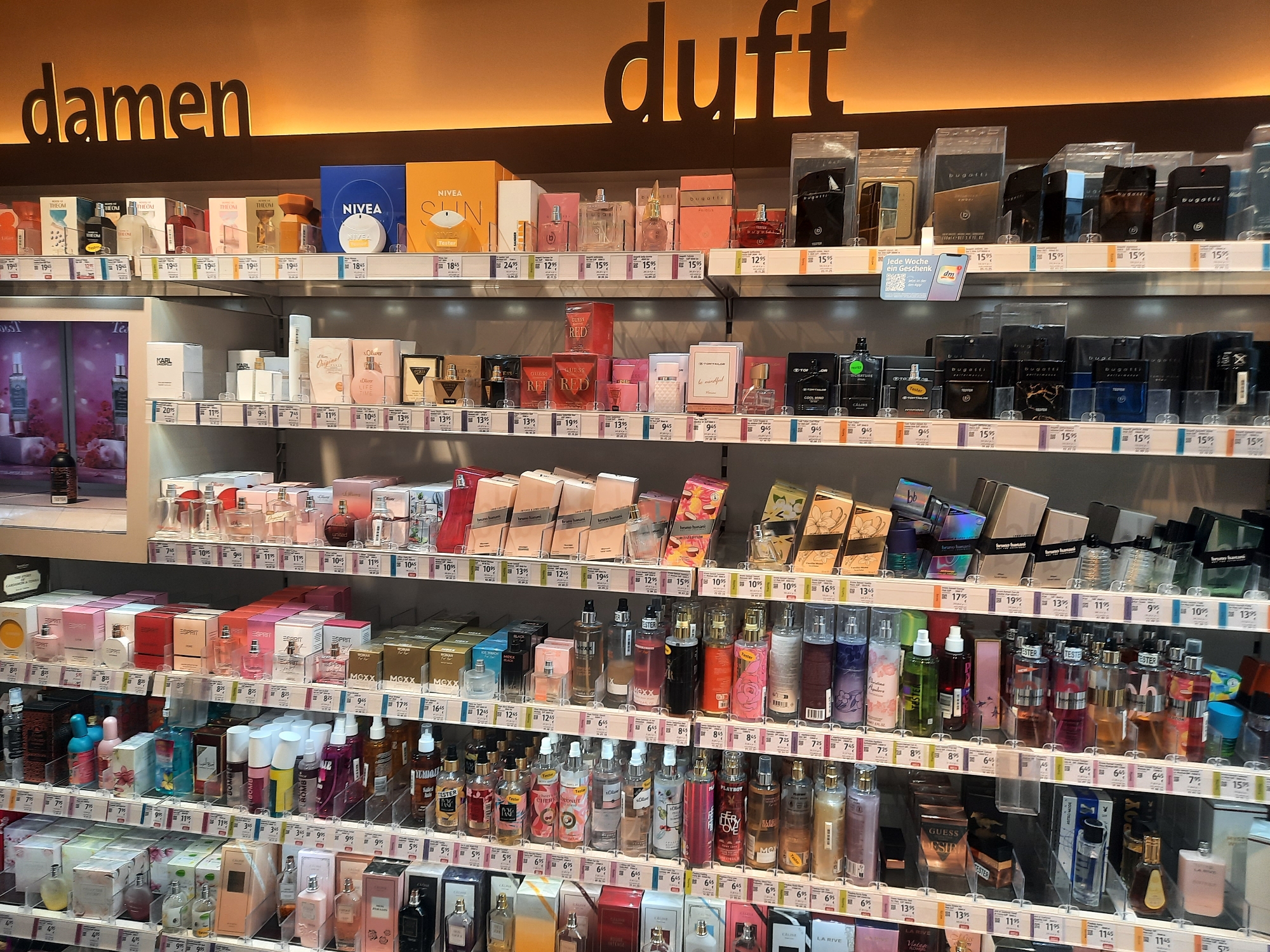 The image shows a well-lit display of perfumes and fragrances in a retail store. The shelves are organized and stocked with a variety of brands and sizes of perfume bottles. Text on the wall above the display indicates "damen" (women) and "duft" (fragrance) in large, dark lettering. The store appears to be a dm-drogerie markt, a common drugstore chain in Germany. The specific location is identified as a dm store in Rosenheim, Germany, based on the presence of the dm logo and the language on the signage. There are no people visible in the image, and the focus is entirely on the merchandise. The products are neatly arranged, with price tags visible below many of the items, indicating a commercial setting. The overall impression is one of an organized and abundant selection of beauty and personal care products.