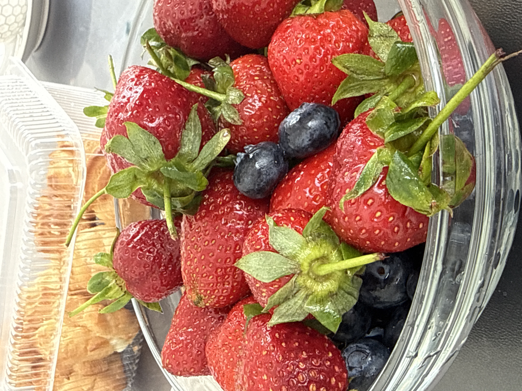 A close-up shot depicts a clear plastic container filled with strawberries and blueberries, positioned near a partially visible plastic container containing baked goods. The berries appear fresh and ripe. The scene appears to be indoors, possibly in a kitchen or dining area. There is no information to confirm this image was taken in the city of Olginskaya, Russia.