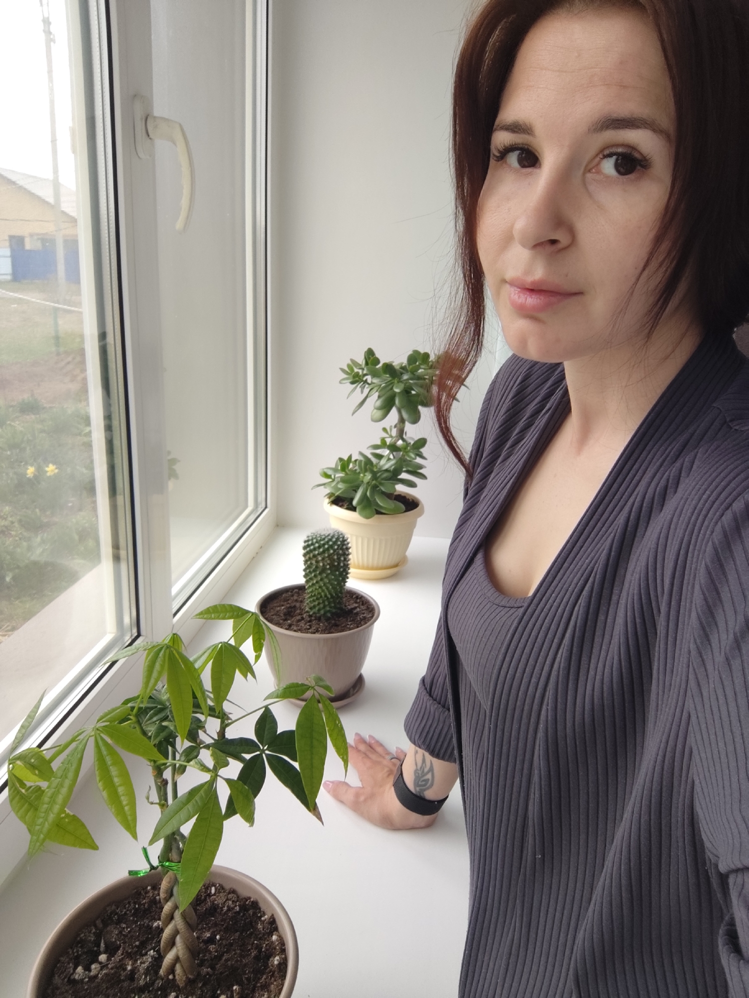 A woman with dark hair stands next to a window with several potted plants on the windowsill. The plants include what appears to be a money tree, a cactus, and two jade plants. Outside the window, a landscape with a building and greenery is visible. The woman is wearing a gray ribbed outfit, and there is a tattoo on her wrist. The location is Фоки, Russia.